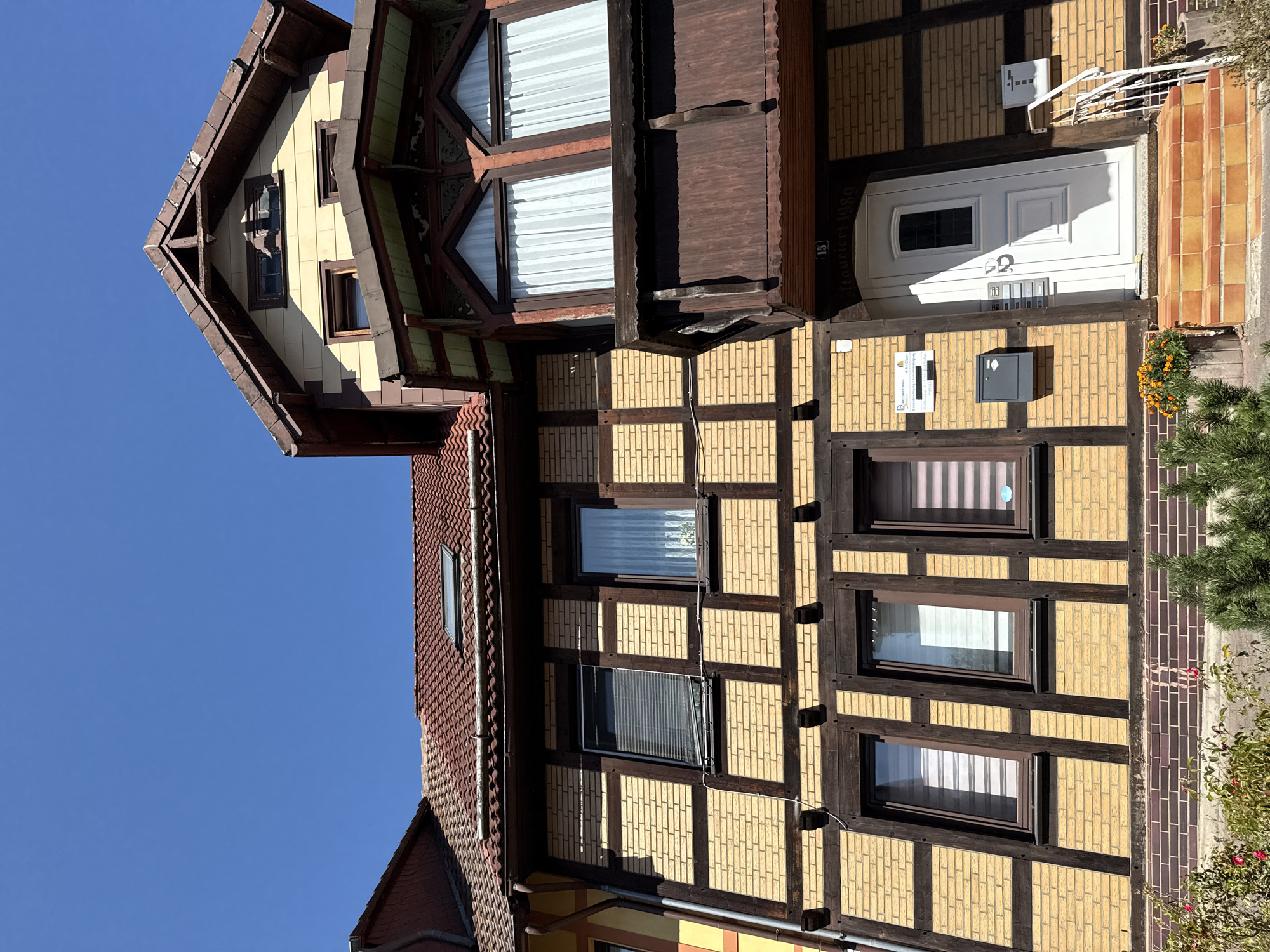 The image shows the facade of a timber-framed house in Bad Sachsa, Germany. The house has yellow brickwork with dark brown timber framing. There are several windows on different levels, some with blinds and others with white curtains. A prominent feature is a dark brown wooden balcony on the upper floor, with decorative elements and large, arched windows. The main entrance to the house is a white door with a small window, accessed by a set of tiled steps. There is a mailbox and an intercom system next to the door. The house is adorned with a roof made of red tiles, and a skylight is visible. The sky above is a clear, bright blue. In the foreground, there are planters with flowers and greenery. Above the door, there is a house number "15" and lettering that appears to read "Roucieri 1989".