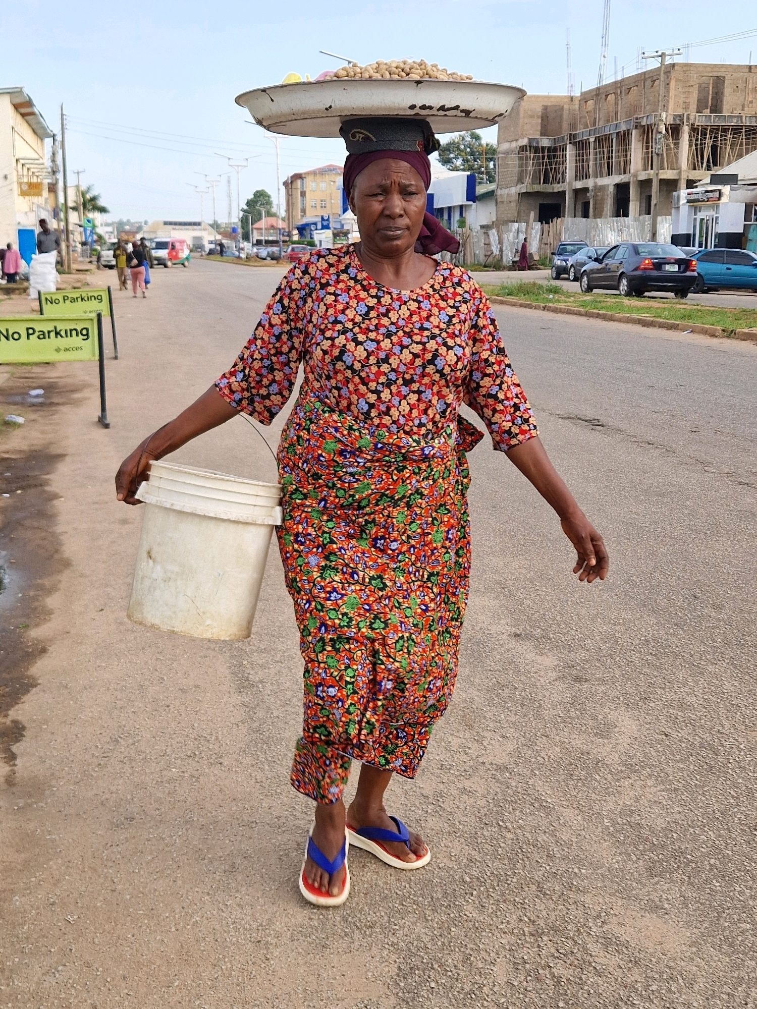 An older woman with dark skin, wearing a vibrantly patterned floral dress and a burgundy headscarf, walks along a paved road in Jos, Nigeria. She carries a large, flat pan filled with nuts balanced on her head and a white plastic bucket in her right hand. The road has markings and is lined with buildings under construction, some with scaffolding, and other more established structures. Several cars are visible on the road, and there are "No Parking" signs on the left side of the street, indicating a commercial or urban area. The sky is overcast.