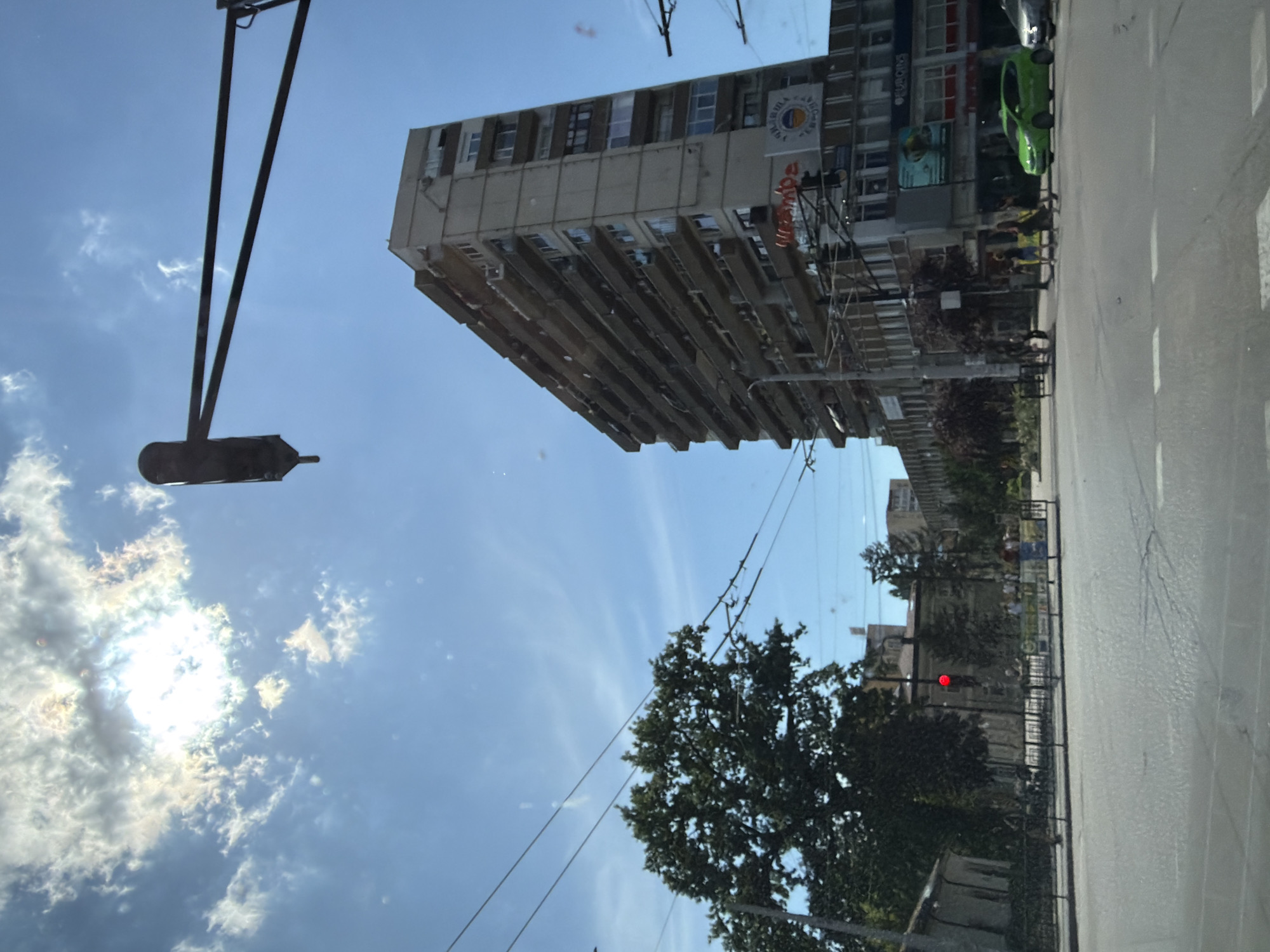 An urban street scene in Pleven, Bulgaria, is captured on a bright, sunny day. The image is taken from what appears to be a moving vehicle, looking upwards and across the street. A multi-story building dominates the left side of the frame, characterized by numerous windows and balconies. Visible on the building are several signs; a circular logo displays text in Cyrillic, including "ПЛЕВЕНСКИ УНИВЕРСИТЕТ" (Pleven University) and "СТОПАНСКИ ФАКУЛТЕТ" (Faculty of Economics), suggesting a university or faculty building. Another sign reads "ФАМИЛА ПАРФЮМЕРИЯ" (Familia Parfumeriya - Family Perfumery), indicating mixed-use occupancy. Below the university logo, orange text, possibly "adepol," is also visible.
The sky above is a clear blue with scattered white clouds, some of which are brightly illuminated by the sun, causing a lens flare effect. A long, dark streetlight pole with an attached fixture extends into the sky from the left foreground. Power lines crisscross the upper and middle portions of the image.
On the right side, a paved street or road runs parallel to the bottom of the frame, with white dashed road markings visible. A bright green car is parked or driving near the building's base. Several small, indistinct figures appear on the sidewalk, suggesting pedestrian activity. A lush green tree stands near the street in the lower middle. Further down the street, a traffic light displays a red signal. The overall atmosphere is that of a bustling city environment during daylight hours.