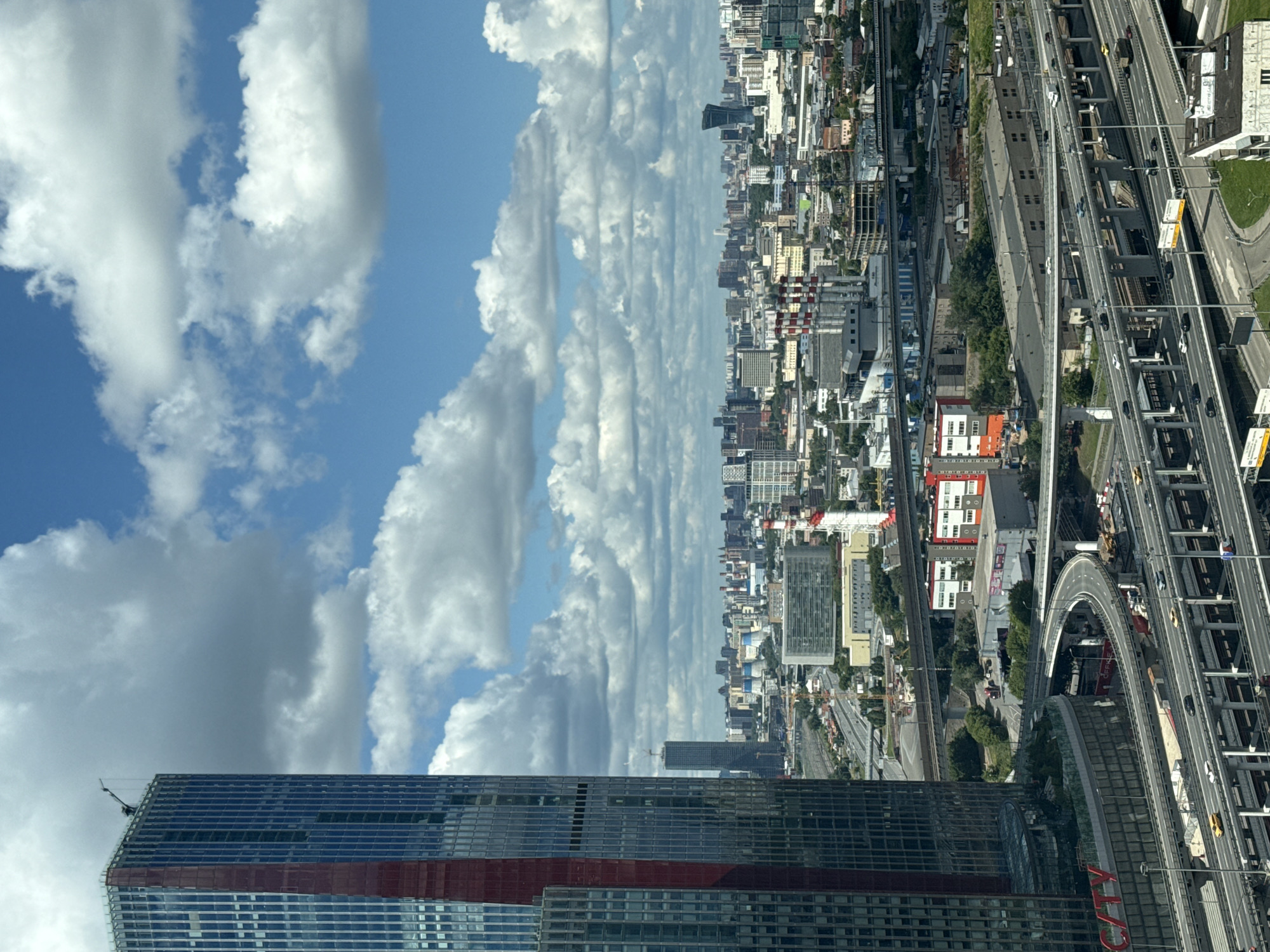 An aerial, eye-level, wide shot captures a sprawling cityscape under a dynamic sky filled with white, cumulus clouds. In the foreground, a towering, modern skyscraper with a blue-tinted glass facade and a vertical red stripe dominates the left side of the frame. A construction crane is visible at the apex of the building. The skyscraper is positioned to the left of a complex network of elevated highways and overpasses, with multiple lanes of traffic visible, including cars and trucks. The highways snake through the urban landscape, dissecting various industrial and commercial buildings. Further into the background, a dense collection of diverse architecture unfolds, featuring residential buildings, office blocks, and what appear to be industrial facilities with chimneys. Distinctive red and white striped chimneys are prominent among these structures. The cityscape extends to the horizon, with a hazy, distant view of more buildings and infrastructure. The overall environment suggests a bustling, developed urban area. The scene is consistent with a view of Khimki, Russia, given the architectural style and the presence of such extensive infrastructure.