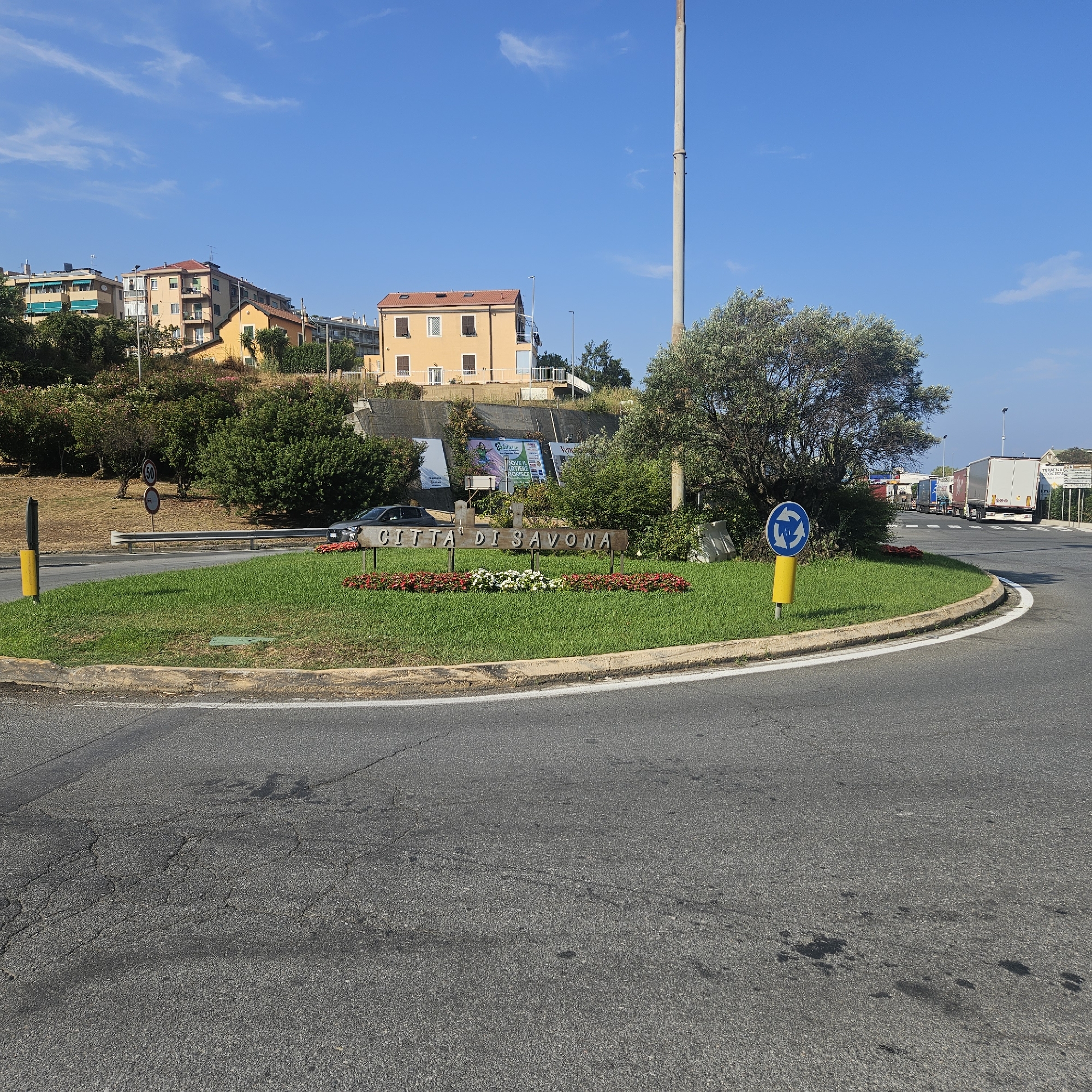 The image depicts a sunny day in the city of Savona, Italy, specifically at a roundabout. In the foreground, the roundabout is covered in lush green grass and decorated with a bed of red and white flowers. A wooden sign with the inscription "CITTA' DI SAVONA" is prominently displayed in the center, signifying the entrance to the city.
To the left, a black car is parked near the sign. Further back, a series of buildings with orange and yellow facades are visible on a hillside, interspersed with trees and greenery. A tall, slender industrial smokestack rises into the clear blue sky behind the buildings.
On the right side of the roundabout, a traffic sign indicates a roundabout and a directional arrow. Beyond the roundabout, a road leads towards a line of parked trucks, suggesting proximity to a port or industrial area. The road surface is asphalt and shows signs of wear with some cracks and dark stains. The overall environment is a blend of urban landscape and nature, with a clear sky and bright sunlight contributing to a pleasant atmosphere.