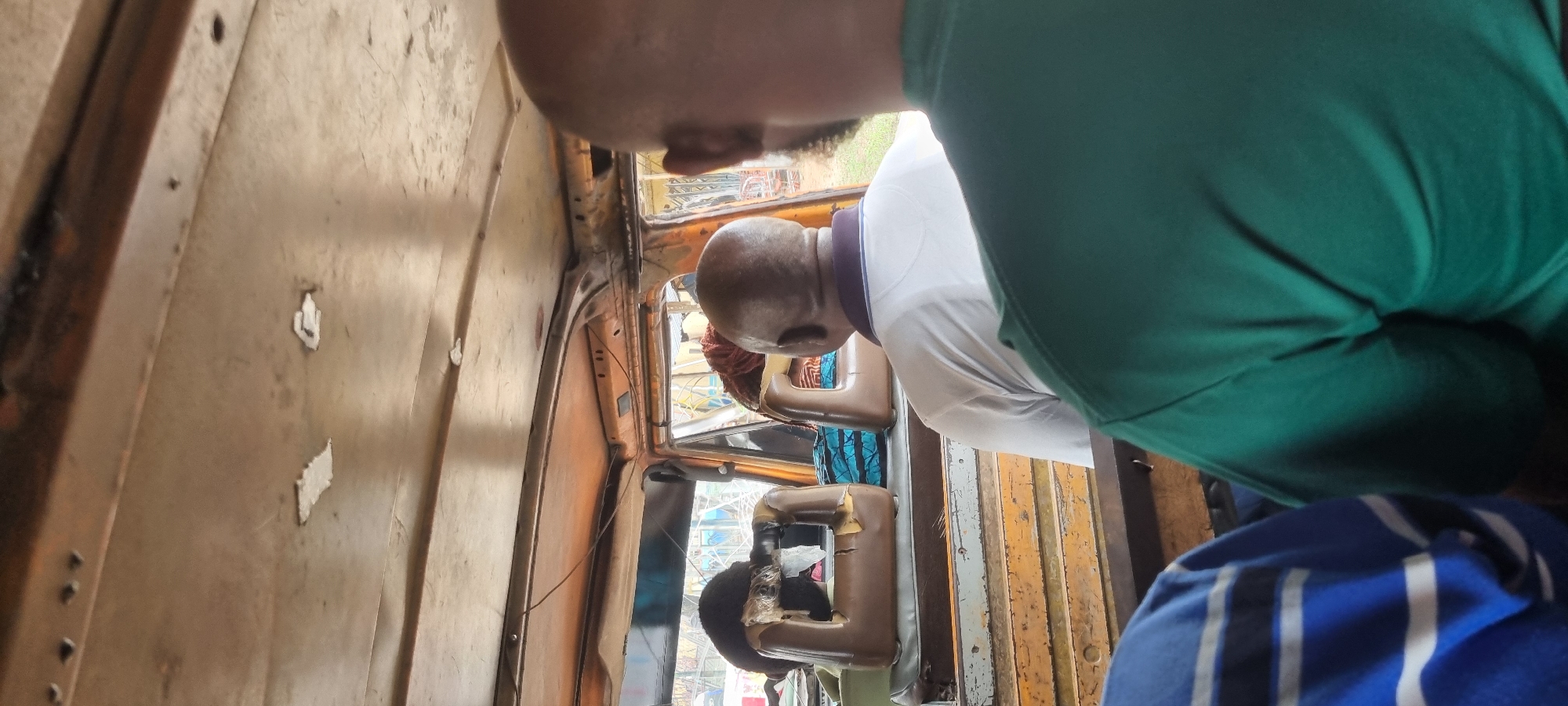 The image shows the interior of what appears to be a crowded motorized rickshaw or tuk-tuk, likely in Ikeja, Nigeria. In the foreground, the back of a person wearing a green t-shirt is visible, with another person wearing a blue and white striped shirt to their left. In the middle ground, a man with a shaved head wearing a white t-shirt with a purple collar is seated, facing forward. Behind him, two worn, brown plastic seats are visible, one with a dark object or person on it, partially obscured. The interior of the vehicle is rustic, with exposed metal and orange-colored padding. The ceiling is also metallic and appears worn. Through the open sides and front of the vehicle, a blurry outdoor environment with buildings and possibly some vegetation is visible, suggesting an urban setting. The overall impression is of a busy, utilitarian mode of transport.