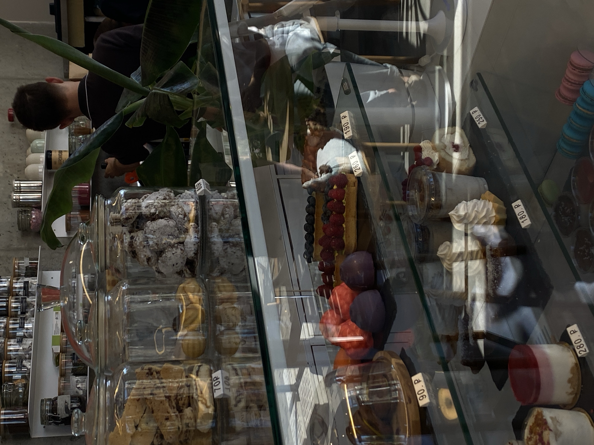 The image depicts a refrigerated display case in a bakery or cafe, likely located in Dinskaya, Russia. The case contains a variety of pastries and desserts. In the foreground, on the bottom shelf, are colorful macarons and what appear to be small cups of dessert. The middle shelf displays several individual cakes and tarts, some topped with berries and cream. One cake is priced at 190 P, another at 250 P, and one at 180 P. Above this, the display case shows more desserts, including some round, plum-colored treats and a tart with raspberries and blueberries, priced at 90 P. Behind the display case, a person, likely an employee, is visible from the back, wearing a dark shirt. The background shows shelves stocked with jars and containers, and a large plant. The overall environment suggests a commercial establishment focused on selling baked goods.