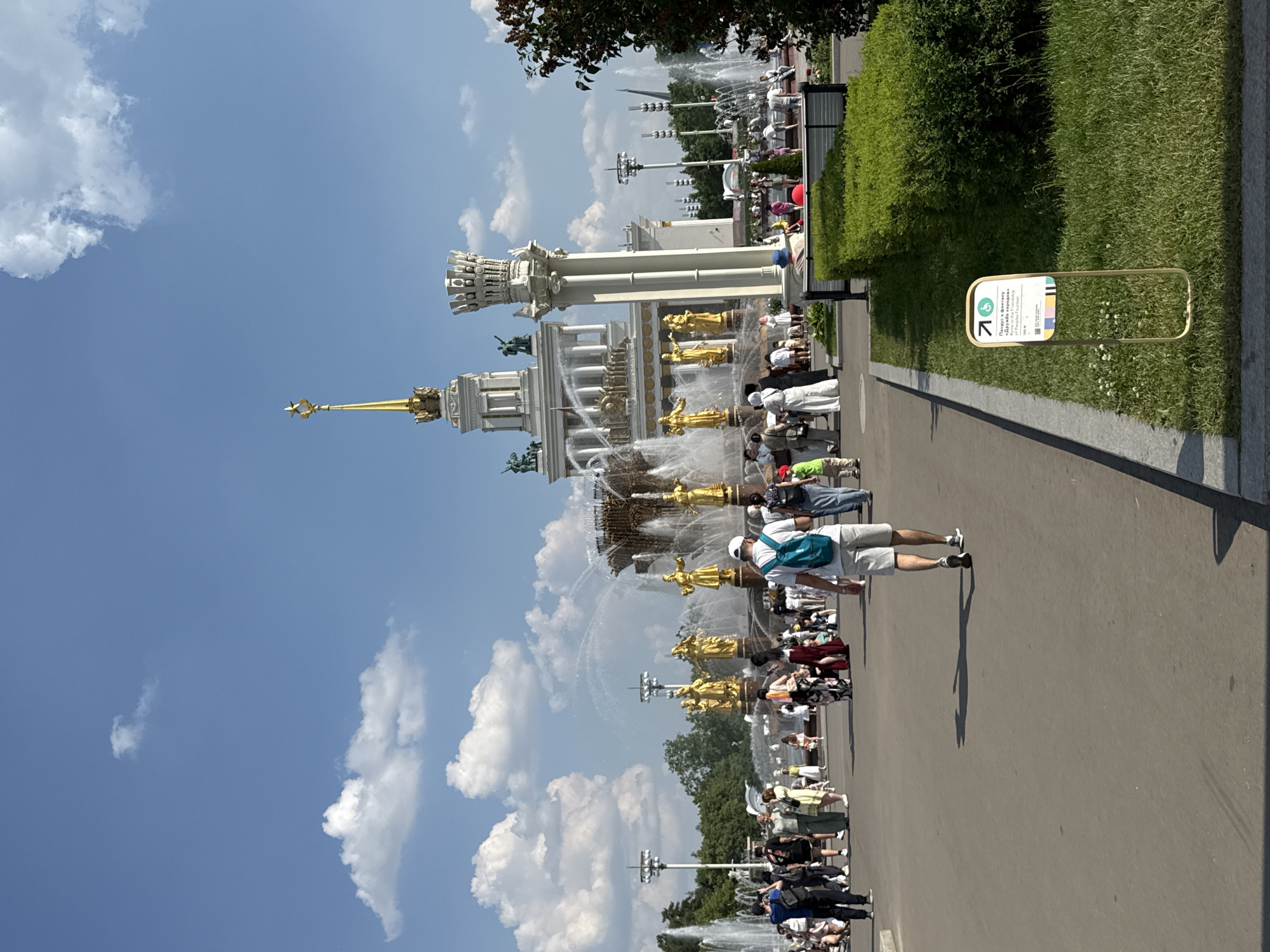 The image depicts the Friendship of Nations Fountain at VDNKh (Exhibition of Achievements of National Economy) in Moscow, Russia. The fountain is a prominent landmark featuring numerous golden statues of individuals representing various Soviet republics. Water jets spray upwards from the fountain's central structure, which is flanked by tall, ornate columns and a large building with a spire topped with a star.
A diverse crowd of people is visible in the foreground and middle ground, walking and gathering around the fountain. They are dressed in casual summer attire. One man in the foreground, facing away from the camera, wears a light blue backpack and gray shorts. A sign with an accessibility symbol and text in Russian and English, likely indicating directions or information about the fountain, is placed in the lower right quadrant of the image on a patch of grass.
The environment is sunny with a clear blue sky dotted with fluffy white clouds. Lush green trees and well-maintained hedges surround the area, indicating it's a pleasant day for outdoor activity. The overall scene captures a bustling atmosphere at a popular tourist attraction in Moscow.