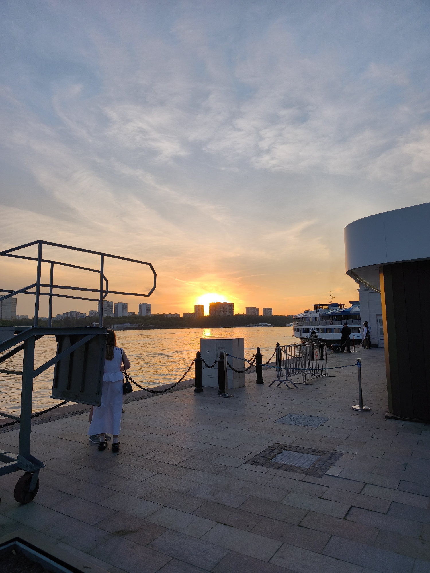 This image captures an urban river embankment in Moscow, Russia, at sunset. The sky transitions from a light blue at the top with wispy cirrus clouds to a vibrant orange and yellow near the horizon, where the sun is visible as a bright orange orb partially obscured by distant buildings.
The Moscow River occupies the midground, its surface reflecting the golden light of the setting sun, appearing orange and yellow with subtle ripples. On the far bank, a line of tall, multi-story city buildings, likely residential or commercial, are silhouetted against the bright horizon, interspersed with a strip of trees below them.
In the immediate foreground on the left, a dark grey metal structure, possibly part of a pier access or maintenance platform, features railings and a visible rusty wheel at its base. The embankment pavement consists of large, rectangular grey and beige stone tiles, with several square and circular manhole covers or grates embedded within. Along the river's edge, dark bollards connected by heavy metal chains delineate the pedestrian area, with temporary light-colored metal crowd control barriers positioned further to the right.
On the right side of the frame, a modern, curved building section, predominantly white with dark grey accents, stands as part of a larger structure, possibly a terminal or restaurant. Adjacent to this building, a large multi-deck riverboat, characterized by a white hull and blue trim, and appearing to have a paddlewheel design, is either docked or moored.
Several people are present on the embankment. A woman with shoulder-length brown hair, wearing a white, tiered dress and carrying a dark handbag, stands with her back mostly to the camera in the mid-left foreground, looking towards the sunset. She wears dark flat shoes. Further to the right, near the riverboat and building, several other figures are visible, including one person in dark attire who appears to be attending to the boat's lines.