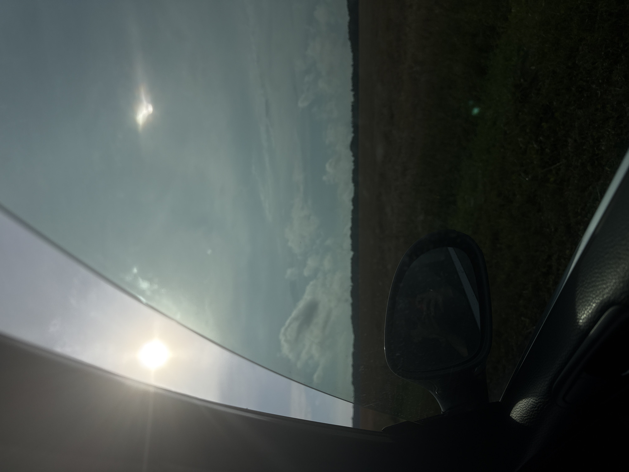 The image captures a view from inside a vehicle looking out the passenger window towards a rural landscape. The sun is visible in the upper left quadrant, partially obscured by the car's window frame. The sky is cloudy, with a second, less distinct solar halo visible in the upper right. A dark field stretches to the horizon, bordered by a line of trees. A car's side mirror is in the lower right portion of the frame, reflecting a portion of the interior of the car and the landscape. The interior of the car, including the door panel, is visible in the lower left. The scene suggests a daytime journey through a flat, open area, possibly farmland or grassland, under a partly cloudy sky. There is no indication of the city of Serya, Russia, or any other specific urban or geographical features.