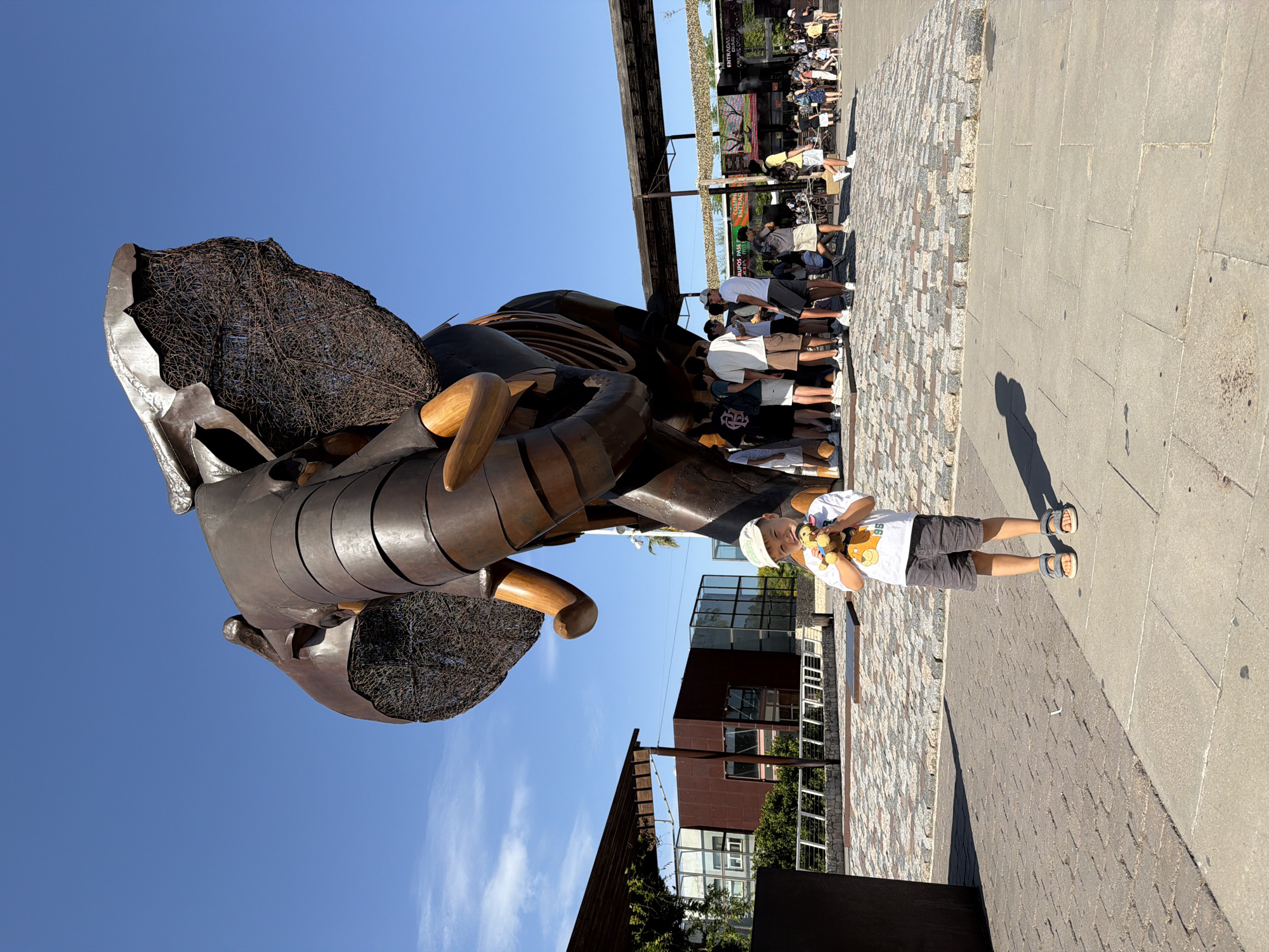 A young boy wearing a white t-shirt, grey shorts, a light-colored cap, and sandals stands in the foreground, holding a stuffed animal. He is positioned in front of a large, dark metal elephant sculpture. The sculpture appears to be made of segmented metal plates and has large, wire-mesh ears. Behind the boy and sculpture, a group of people, mostly young adults, are gathered, and further in the background, there are buildings with modern architecture, some with glass facades. The ground is paved with cobblestones in a patterned design. The sky above is clear and blue. The location is the Valencia Bioparc in Valencia, Spain, known for its large animal sculptures as part of its thematic design.