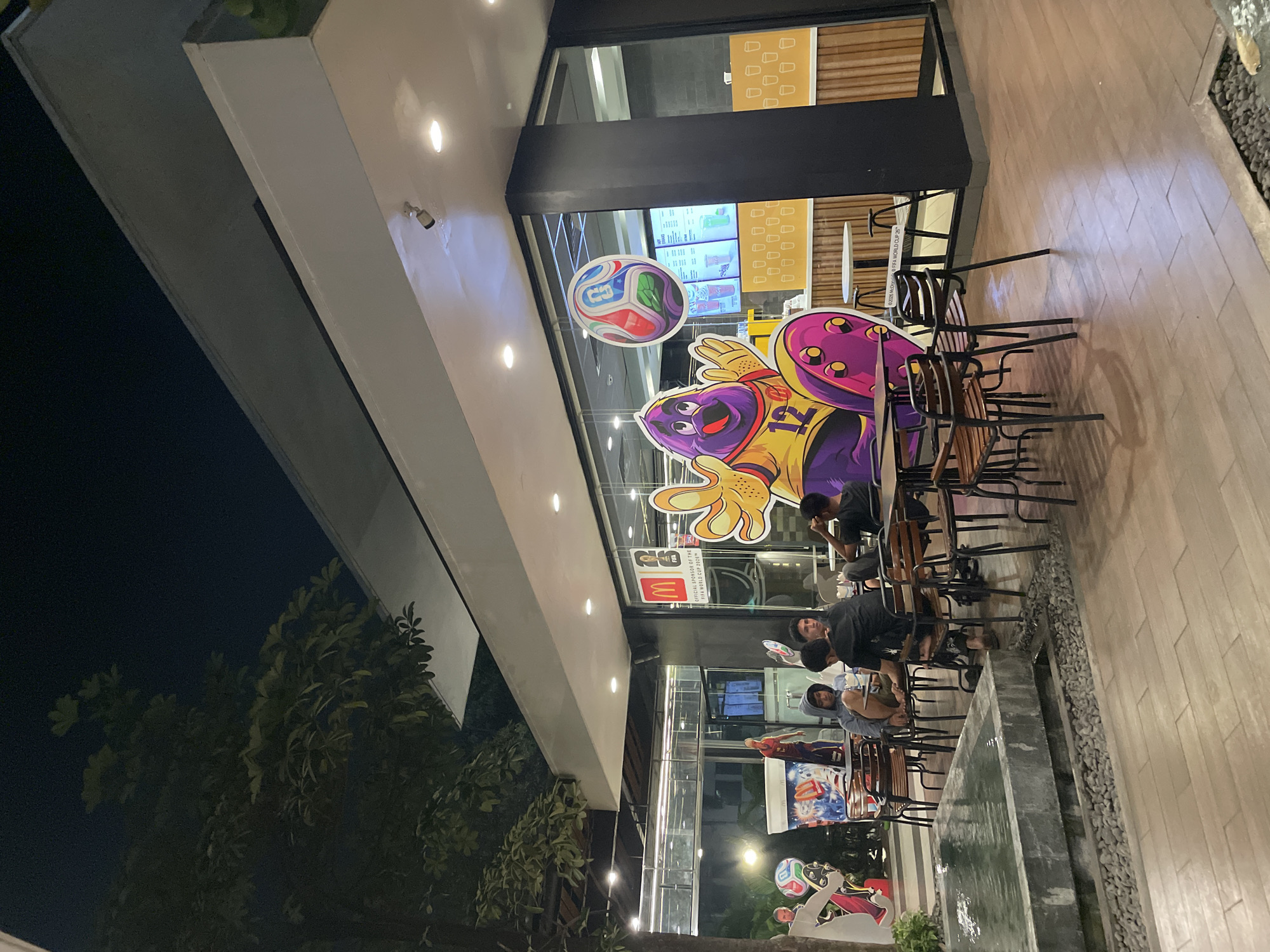 The image shows an outdoor seating area of a McDonald's restaurant at night, located in North Jakarta, Indonesia. Several customers are seated at tables. The restaurant's exterior features a large window display with cartoon characters and a soccer ball graphic, indicating a promotion related to the FIFA World Cup. A small water feature is visible in the foreground. The overall ambiance is casual and illuminated by artificial lighting.
