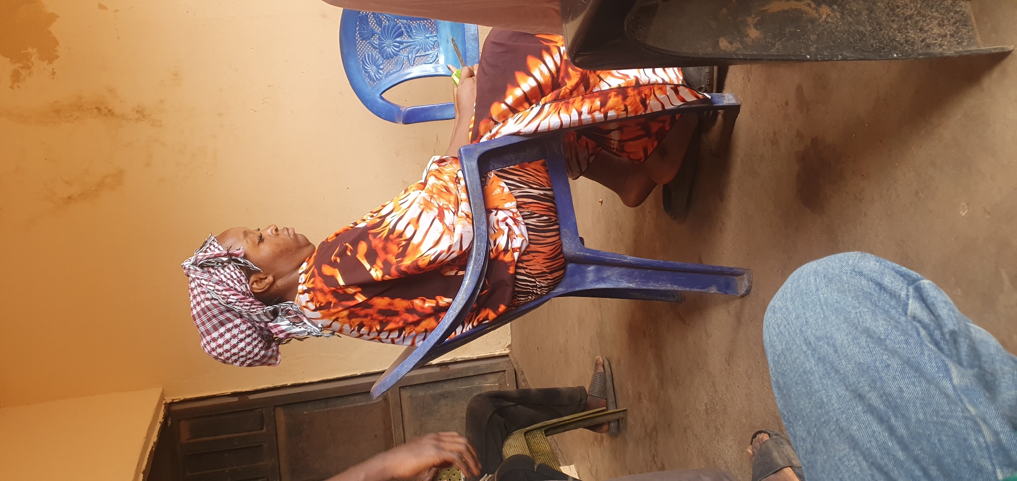 The image captures a candid moment, likely during the daytime, in Idah, Nigeria. The central subject is an adult woman, appearing to be of African descent, seated comfortably in a blue plastic chair. She is dressed in a vibrant, flowing garment featuring an orange, brown, and white abstract pattern, possibly resembling animal stripes. Her head is adorned with a checkered headwrap in red, white, and dark blue/black plaid. Her head is tilted back, resting against the chair, and her eyes appear to be closed or looking upwards, conveying a sense of rest or contemplation.
The setting appears to be an outdoor or semi-outdoor space, characterized by a plain, light brown or tan wall in the background, showing some minor smudges or discoloration. The ground beneath is a similar earthy brown, suggesting dirt or compacted soil. Another blue plastic chair with an ornate backrest is partially visible behind the woman, and a dark, heavy object, possibly a table or furniture, is seen in the upper right.
The presence of other individuals is indicated by partial views of their limbs: a leg clad in blue jeans is visible on the right, and another person's legs in dark trousers with sandals, along with their hands, are seen in the lower left portion of the frame. This suggests a small gathering or social interaction. A dark door or gate is also partially visible towards the bottom left. The overall atmosphere is informal and relaxed. No discernible text is visible in the image.