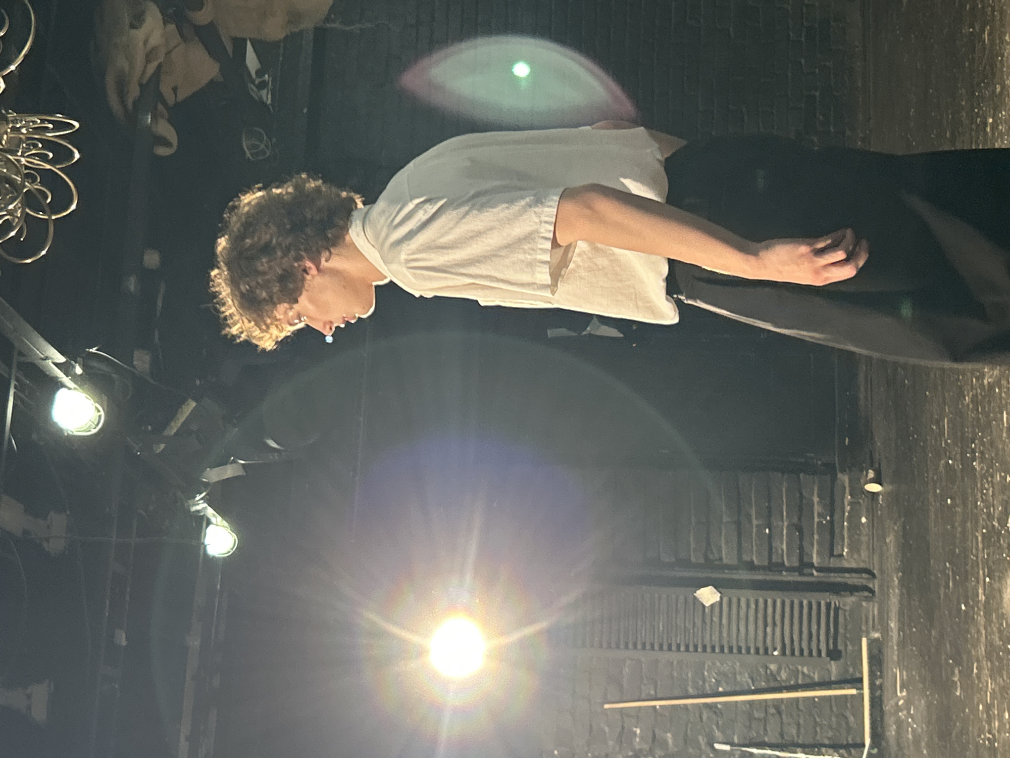 A person with curly brown hair stands facing left, with their head tilted down. They wear a white t-shirt and dark pants. The background is dark, with some stage lighting visible. One light in the foreground appears as a bright, round lens flare, while another overhead light casts a green, oval-shaped glow on the wall behind the person. The scene appears to be indoors, possibly a performance space or studio, given the lighting and dark, brick-like wall. The person's posture and the lighting suggest a moment of contemplation or a pause during an activity. No text is visible.