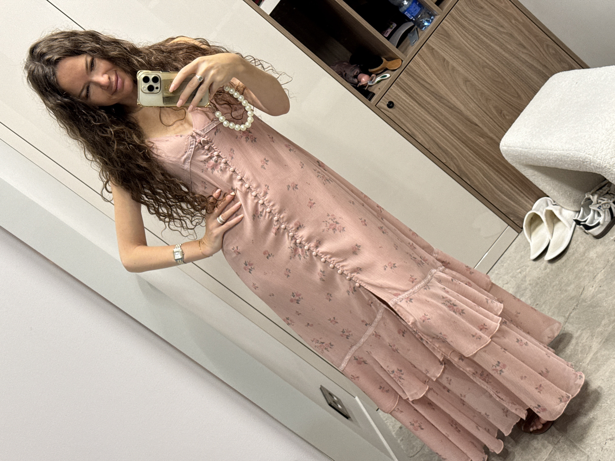 A woman with long, curly brown hair is taking a mirror selfie in a room. She is wearing a long, pink floral print dress with lace trim and a slit up the front. She has on a silver watch and several rings, and a pearl necklace. To her right is a wooden cabinet with shelves containing various items. In the foreground, on the floor, are a pair of white slippers and sneakers. The overall impression is that of a domestic setting. This scene is set in the city of Khimki, Russia.