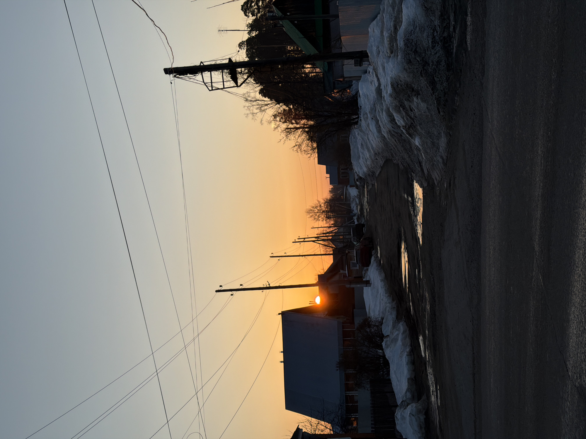 The image depicts a residential street in Chaikovsky, Russia, at sunset or sunrise during late winter or early spring. The sky transitions from a vibrant orange at the horizon, where the sun is partially visible behind a house, to a pale blue overhead.
The unpaved or poorly paved street runs into the distance, appearing dark and wet with several puddles reflecting the orange light of the sky. Accumulations of dirty snow line both sides of the road, indicating melting conditions. On the left, a dark-roofed house with visible windows is silhouetted against the bright horizon, bordered by a dark wooden fence and a small bush. A series of utility poles with overhead wires run along this side of the street.
On the right, larger piles of snow are present. Beyond the snow, several bare deciduous trees and bushes are visible. Further down the street, another house with a dark green roof is partially obscured by trees. The background on the right is dominated by a dense stand of dark coniferous trees, likely a pine forest. A prominent, tall utility pole with complex wiring and a metal frame structure stands on the right side of the foreground, also silhouetted. A red passenger car is parked on the right side of the street further back. No individuals are present or engaged in any discernible actions or interactions.