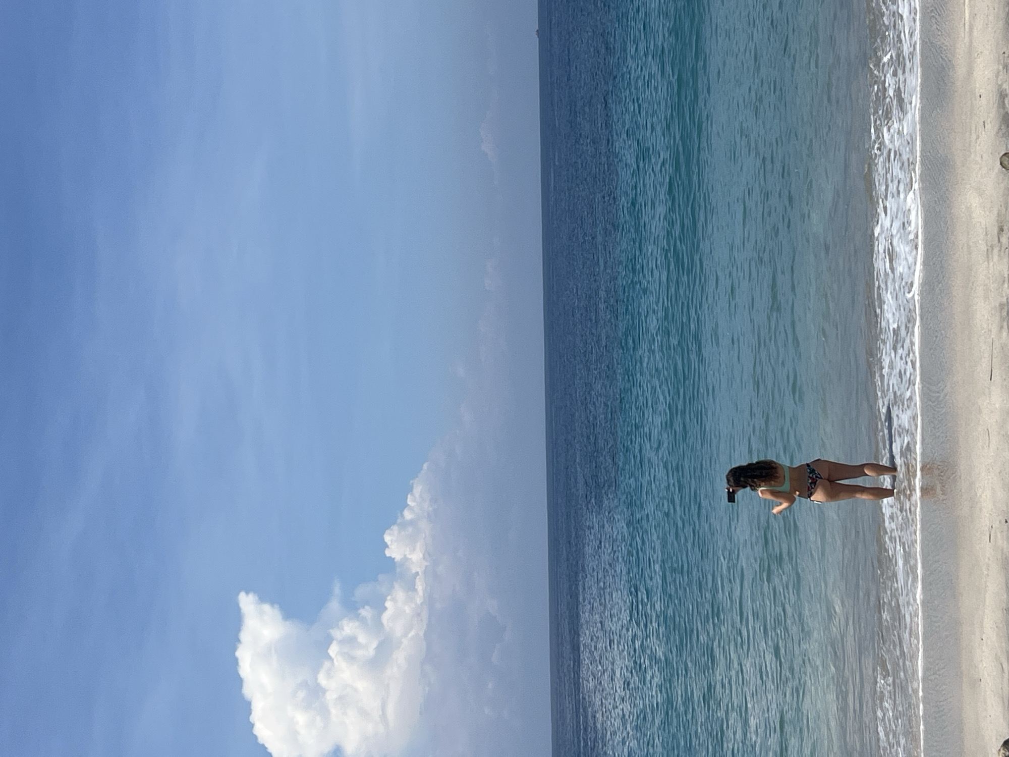 A medium shot, rotated 90 degrees, captures a woman standing at the edge of a beach, waist-deep in the surf. She is holding a mobile phone in her left hand, seemingly taking a photo. She is wearing a bikini in floral patterns, her hair is black, and is pulled back. The water is shallow, turquoise in hue. Behind her lies a wide expanse of ocean, mirroring the clear blue sky overhead. White clouds are visible in the sky. The scene is Karangasem, Indonesia.