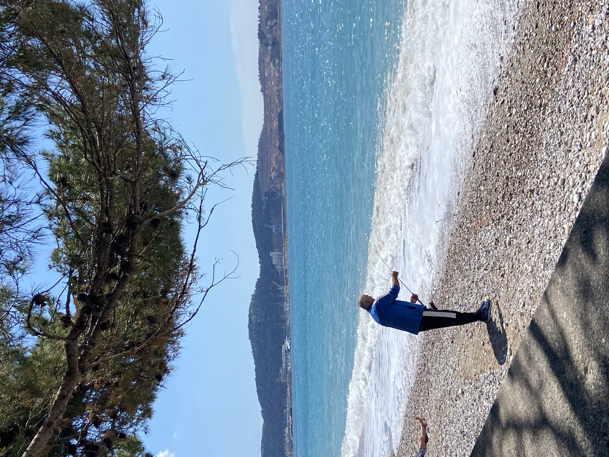 The image shows a man fishing on a pebble beach in Bar, Montenegro. The man is wearing a blue jacket, black pants with white stripes, and sneakers. He is holding a fishing rod and appears to be casting his line into the turquoise water. Waves are washing onto the shore. In the background, a coastal landscape with mountains and buildings is visible under a clear blue sky. To the left, a tree with pine cones partially frames the image. Shadows are cast on a concrete structure in the lower right corner.