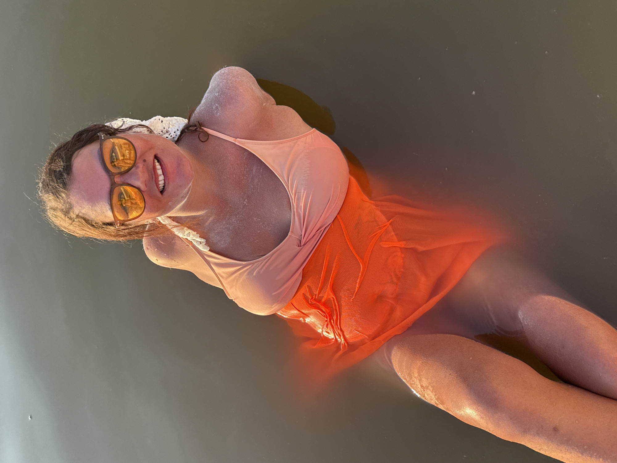 In the city of Sol-Iletsk, Russia, a woman is submerged in murky water, her skin appearing flushed and possibly sunburnt. She is wearing a light pink bikini top and an orange sheer cover-up. She has on orange-tinted sunglasses and is smiling. Her hair is brown and appears wet. The water is dark and still, with some ripples visible. The lighting suggests it is daytime.