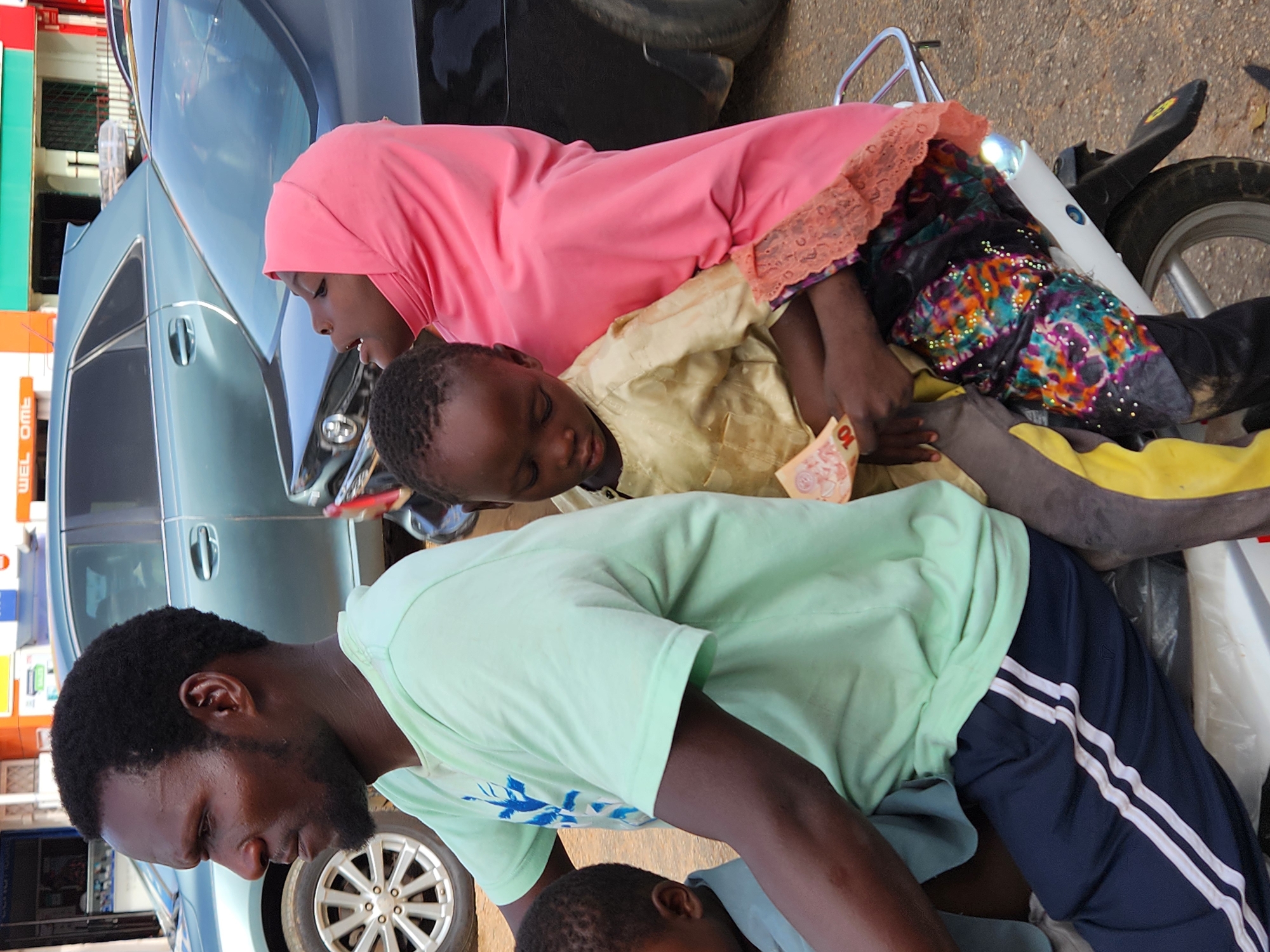 The image shows a man, a woman, and two children in an outdoor setting, likely a city street. The man, in the foreground, wears a light green t-shirt and dark blue track pants with white stripes. He has dark skin, short black hair, and a beard. Above him, a woman is lying on a white object, possibly the seat of a motorcycle. She is wearing a pink headscarf and a colorful, patterned dress. A child lies next to her, wearing a light yellow shirt and holding a banknote. The woman seems to be holding the child. There is also another child, only partially visible, near the man. A blue car is partially visible in the background, along with a building. The exact location within the city is unknown.