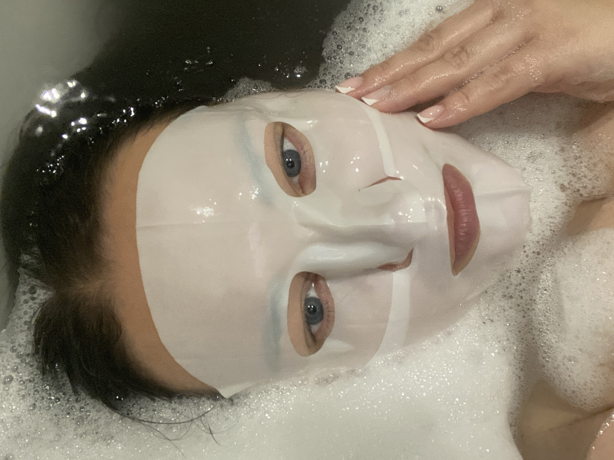 A woman lies in a bath of foamy water wearing a sheet mask. Her dark hair is wet. Her right hand rests on the side of her face. The location is not identifiable as Sochi, Russia based on the image.