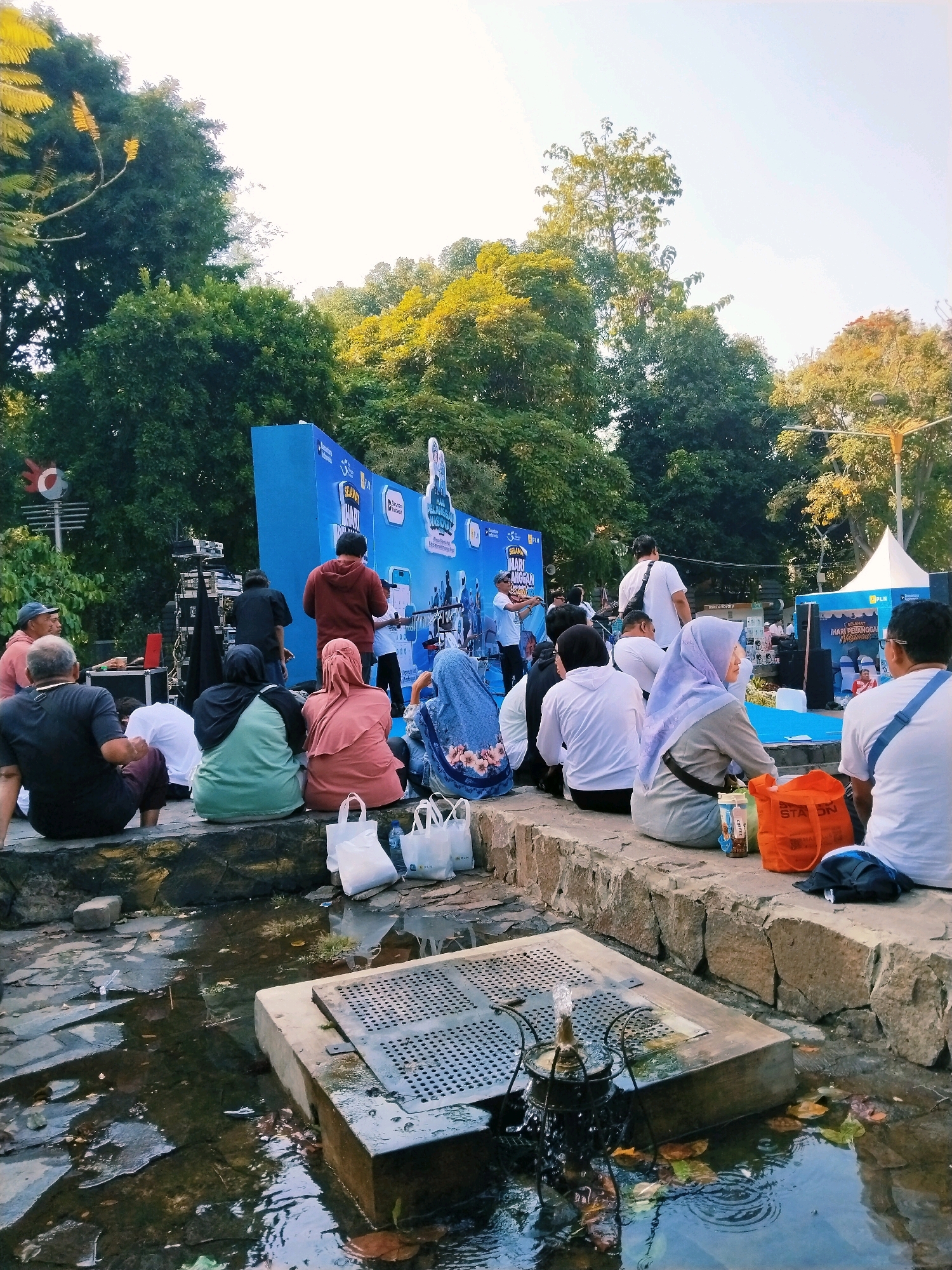 The image depicts an outdoor event in a park-like setting, likely in Surabaya, Indonesia, given the event's banner which reads "SELAMAT JALAN MARI BERSANTAI" and the presence of "PLN" branding, a major Indonesian state-owned electricity company. A group of people are seated on a stone wall, facing a stage area. The stage features a large blue backdrop with event graphics and logos. On the stage, several individuals are present, including performers on a platform and technical crew behind equipment. The audience, composed of individuals of various ages, are casually dressed and appear to be observing the event. Several white plastic shopping bags are placed on the wall in front of the seated audience. In the foreground, a small fountain is visible in a shallow pool of water, with fallen leaves scattered on the surface. Lush green trees and foliage surround the scene, indicating a public park or outdoor recreational area. The lighting suggests it is late afternoon.
