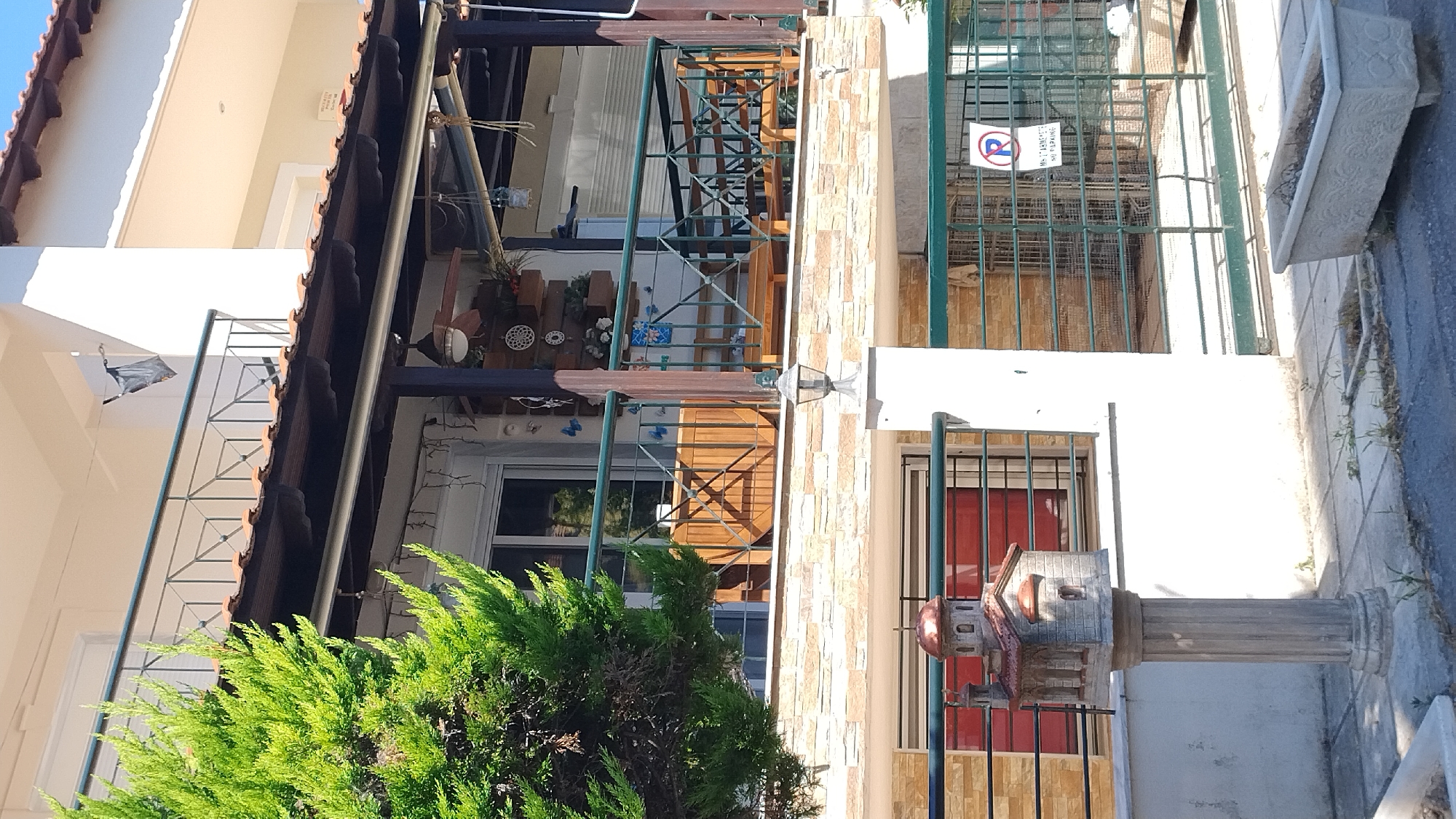 The image captures a sunlit scene in Neoi Epivates, Greece, focusing on the exterior of a building. In the foreground, a decorative concrete column supports a miniature model of a church with a copper-colored roof. Behind this, a railing with green metal bars runs horizontally, separating the ground level from the upper floor. A window with a red-tinted glass is visible on the left.
The building's facade is adorned with light-colored, textured stone blocks. Above this, a balcony features a dark-colored railing with a diamond pattern and a wooden overhang supported by dark posts. The balcony itself contains outdoor furniture, including a wooden table and benches. A large green conifer tree partially obscures the left side of the building.
Further back, a green metal fence encloses an area, and a circular sign with a red 'R' inside, along with Greek text indicating "NO PARKING," is attached to it. A concrete planter box sits on the ground in front of the fence. The sky above is clear and blue.