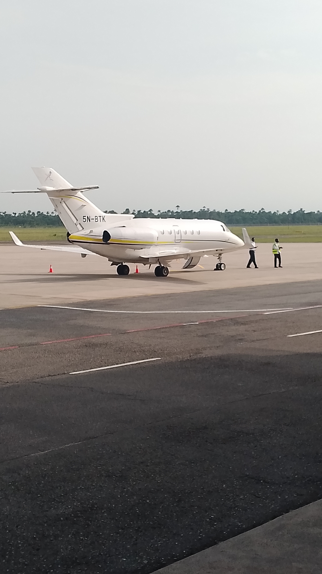 The image depicts a white private jet with registration number 5N-BTK parked on an asphalt runway. Two individuals are standing near the front of the aircraft. In the background, a row of trees is visible, with an overcast sky above. The location is presumed to be in or near Orerokpe, Nigeria. The ground is asphalt and there are white and red runway markings. There are orange traffic cones beside the jet.