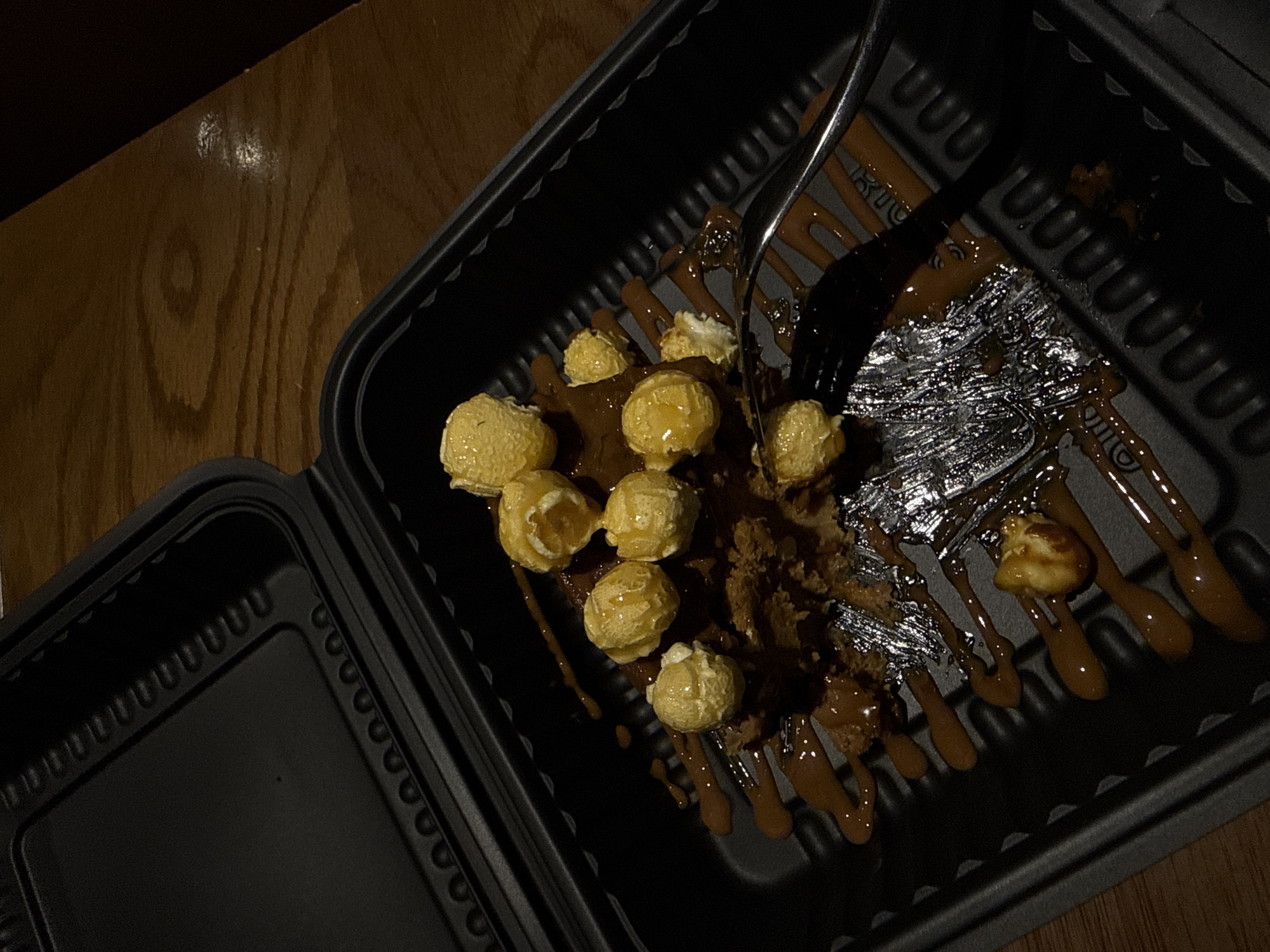 The image shows a black clamshell container on a wooden surface. Inside the container is a partially eaten dessert consisting of a dark base, topped with popcorn and a caramel-like sauce. A metal fork is piercing the dessert. The surrounding environment is dimly lit. There is no verifiable information to place the location of the photo in San Bernardino Tlaxcalancingo, Mexico.