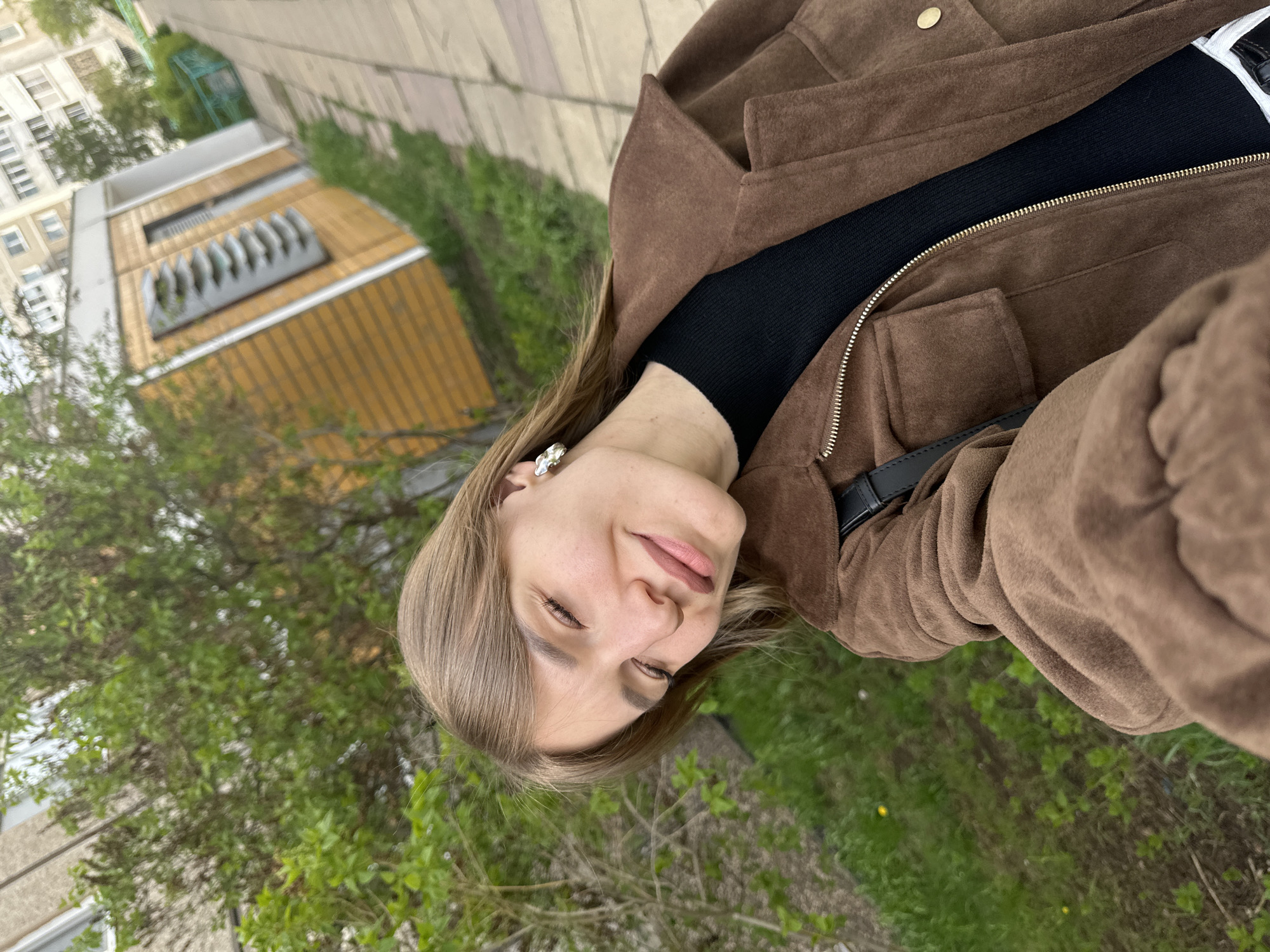 A selfie image shows a young woman with fair skin, light brown hair, and closed eyes. She is wearing a brown suede jacket over a black top. An earring is visible in her left ear. The background includes green foliage and a building with a yellow facade and horizontal detailing, potentially located in the city of Chaikovsky, Russia. A sidewalk or paved area is visible at the top of the image.