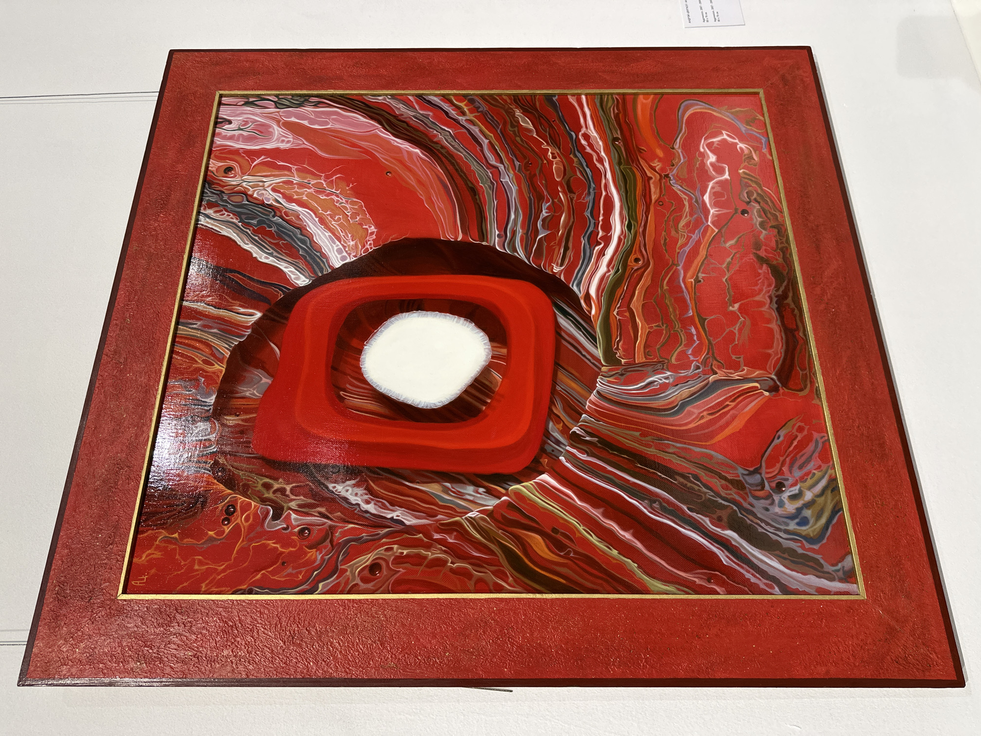 The media file displays an abstract artwork, likely a painting, predominantly featuring various shades of red, alongside swirling patterns of white, orange, black, gray, and subtle hints of gold or bronze. The artwork is set within a textured, matte red frame, delineated from the painted surface by a thin, golden inner border.
The central composition includes a dark red, almost oval shape, from which a brighter, rounded square emerges. Within this square lies a prominent, irregular white, circular form, acting as a focal point. Surrounding these central elements, the painting exhibits dynamic, flowing patterns, characteristic of fluid art or acrylic pouring techniques. These patterns create organic, marbled lines and cellular formations across the canvas, showcasing a glossy finish that reflects light.
The artwork is presented against a plain white wall, suggesting its display in an art gallery or exhibition setting. A small white label with black text is partially visible in the upper right background of the original image. While most of the text is illegible due to distance and angle, some discernible Cyrillic characters and numbers are present, hinting at artwork details such as "см" (centimeters) for dimensions, and a potential date range like "2020-2021". There are no people or specific events depicted; the scene is static, focused entirely on the artwork itself.