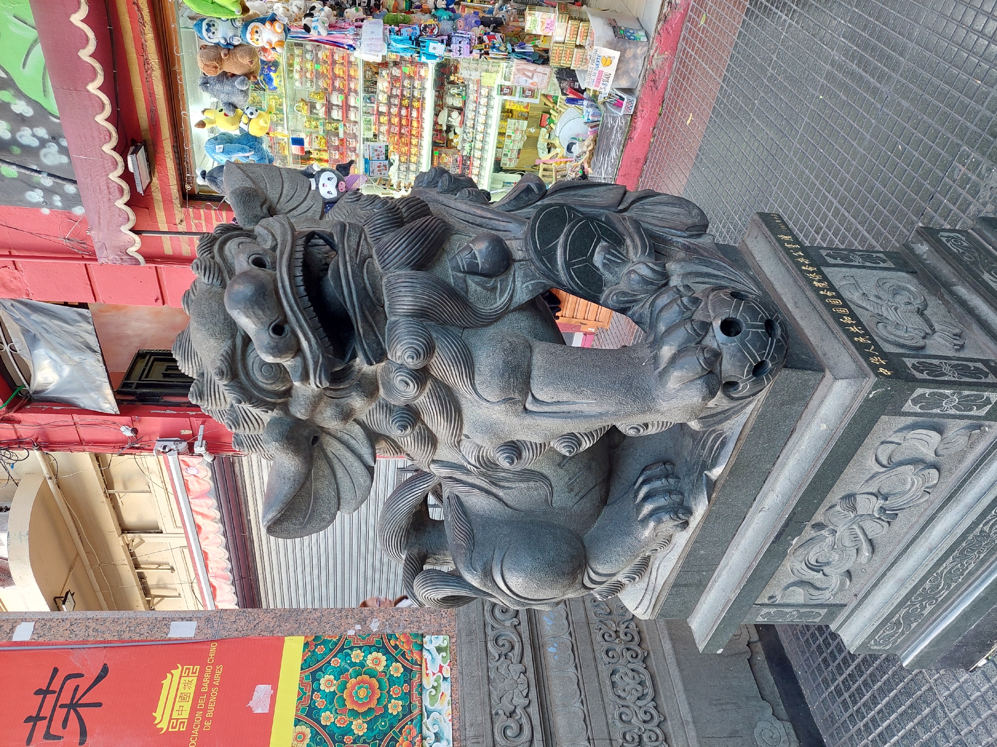 The image captures a detailed stone statue of a Chinese guardian lion (Foo dog) prominently in the foreground, angled to the right. The dark gray statue features intricate carvings of its mane, fierce expression, and claws, with one paw resting on a spherical object. Its pedestal is also carved with decorative patterns and Chinese characters.
Behind the statue, a vibrant commercial street scene unfolds. To the left, a storefront with a red awning displays a large red banner. The banner features Chinese characters "荣" and "中國城記", along with the Spanish text "ASOCIACION DEL BARRIO CHINO DE BUENOS AIRES", clearly identifying the location as the Chinatown of Buenos Aires, Argentina. A colorful floral mural is visible next to the banner.
To the right of the statue, another brightly lit shop window showcases a variety of merchandise, including numerous plush toys (some resembling cartoon characters) and other small items. A yellow sign in the window reads "SUERTE" (Luck) and "$3.000".
The base of the guardian lion statue also bears an inscription in Chinese, which translates to "Presented by the Overseas Chinese Affairs Office of the State Council of the People's Republic of China." The ground is paved with textured tiles. The bright lighting suggests it is daytime, highlighting the cultural elements and commercial activity of the neighborhood.