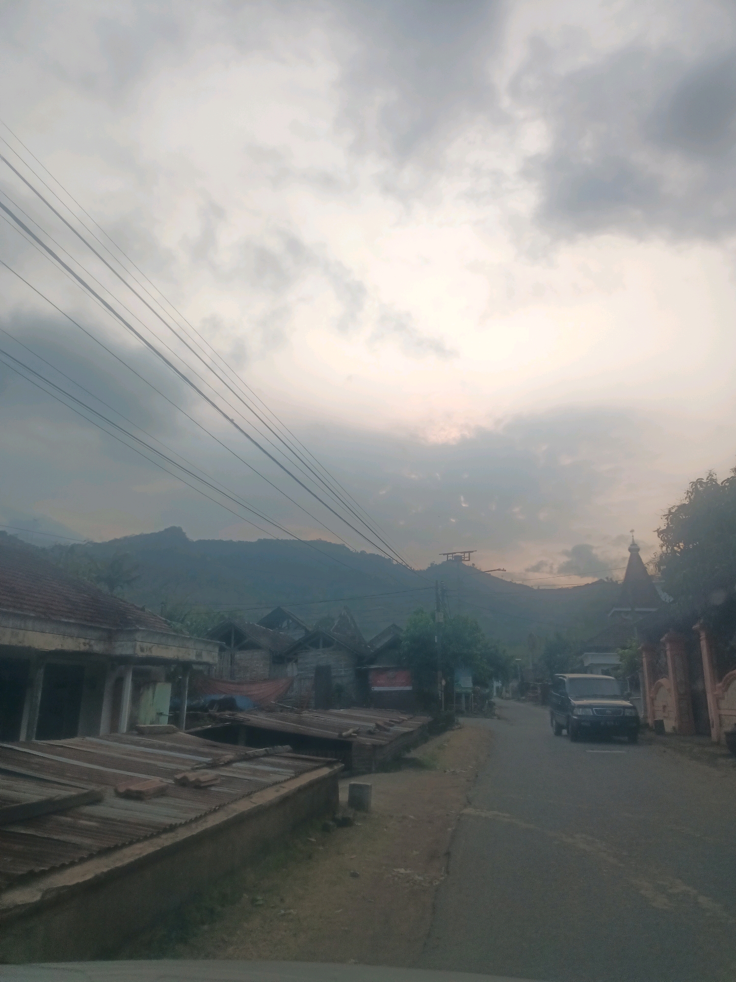 The image captures a street scene in the city of Gandusari, Indonesia, characterized by a cloudy sky and a misty, diffused light, suggesting either early morning or late afternoon. The foreground is dominated by the edge of a road and a pathway, with several low-lying buildings and structures lining the left side. These buildings appear to have corrugated metal roofs, some in disrepair, and are constructed with simple materials, typical of a rural or semi-urban Indonesian setting.
Further down the road, a dark-colored SUV is visible, driving away from the camera. The road itself is paved but appears worn. On the right side of the road, a decorative fence and a building with a pointed roof, possibly a small religious structure or a uniquely designed house, are present. In the background, rolling hills or mountains are shrouded in a hazy atmosphere, blending with the cloudy sky. Overhead, multiple electrical wires stretch across the frame, adding a sense of infrastructure to the rural landscape. The overall impression is one of a quiet, overcast day in a modestly developed area of Gandusari.