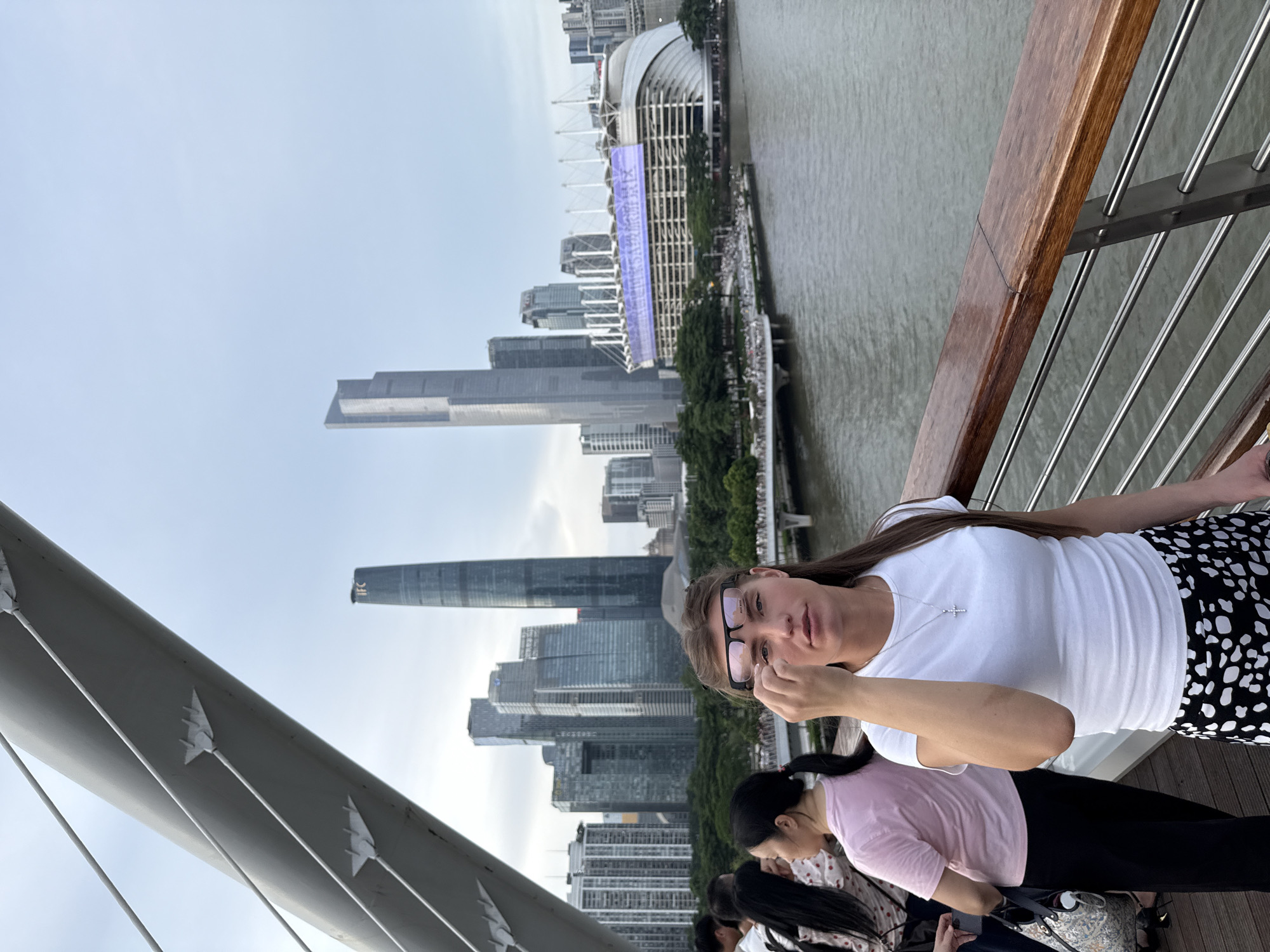 A woman with long brown hair and glasses is in the foreground, looking at the camera. She is wearing a white t-shirt and a black and white spotted skirt. The woman is standing on a bridge, with a cityscape and a river in the background. Other people are visible on the bridge and in the distance. The sky is overcast, suggesting it is either early morning or late afternoon. The overall scene appears to be a busy urban environment, possibly a tourist area. There is no visible text in the image.