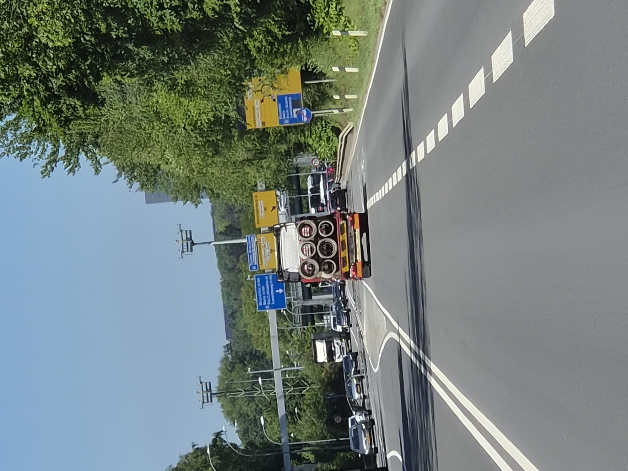 A slightly overexposed, mid-level, medium-distance shot depicts a northbound highway in Luxembourg. A white, long-bed semi-truck is traveling in the right lane; it's carrying six large metal cylinders. The left lane is occupied by a queue of passenger vehicles of mixed makes and models. Yellow directional road signs are visible over the median barrier, positioned alongside streetlights. Heavy foliage and trees line the left side of the highway. The sky is mostly clear with a pale blue hue. White lane markings are clearly visible on the road surface.