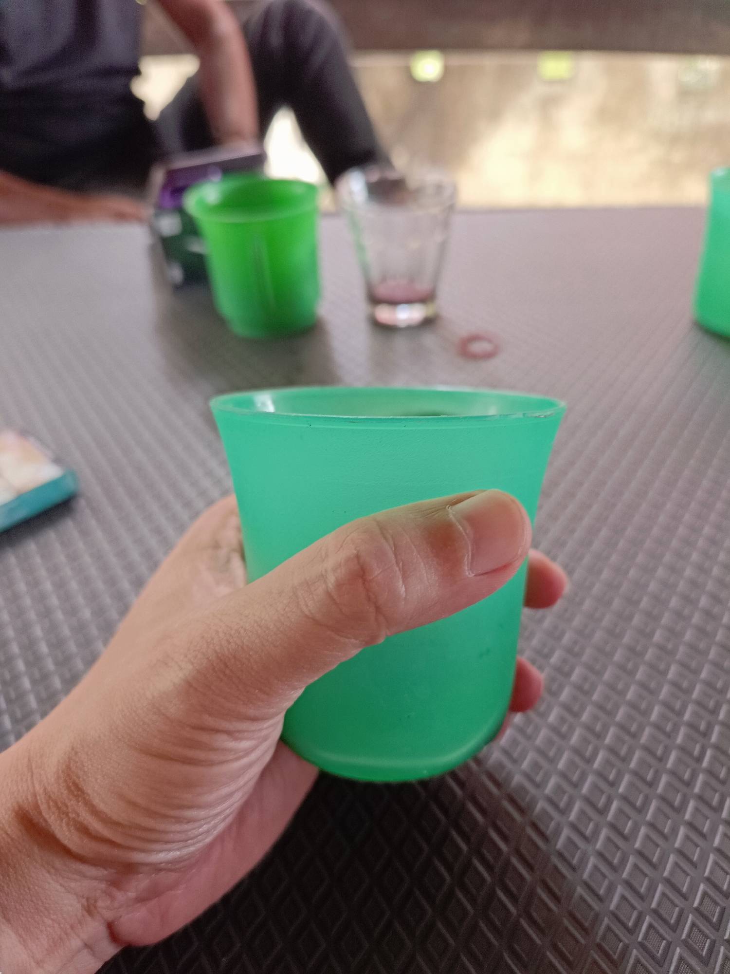 A hand holds a bright green plastic cup on a textured grey table. In the background, out of focus, are two other cups, one green and one clear glass, and the lower body of two people. The person in the foreground appears to be wearing dark pants, and the person in the background is wearing a dark shirt. The environment seems to be outdoors, possibly at a cafe or rest stop, with a blurred background suggesting a wall and natural light.