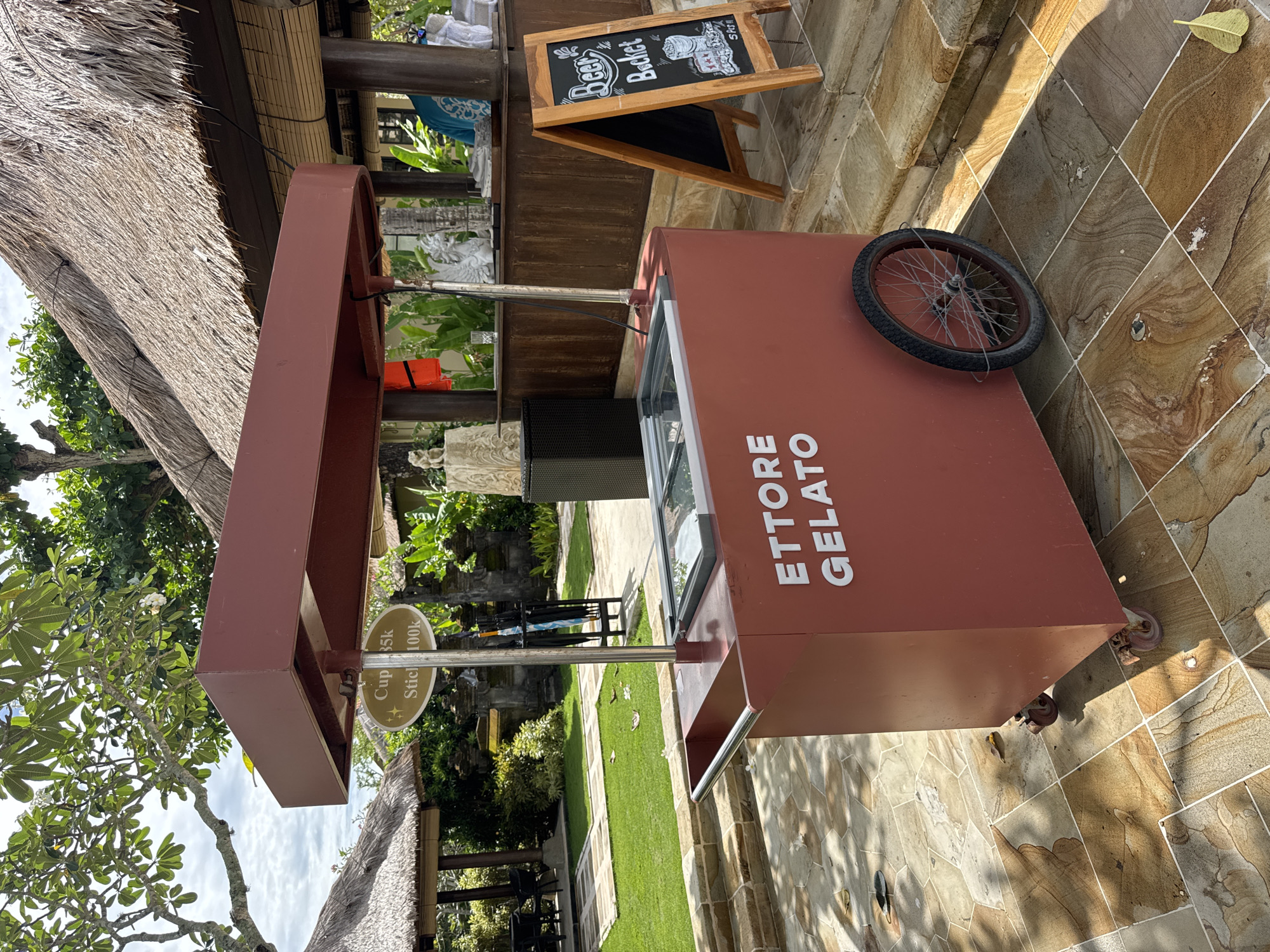 The image shows an "Ettore Gelato" cart with a wheel, located outdoors in a paved area. Behind the cart, there is a wooden structure with a chalkboard sign that reads "Beer Bucket". The surrounding environment includes greenery, suggesting a garden or park setting. There is a sign that says "Cup 35k Stick 100k". The location is in Kuta Selatan, Indonesia.