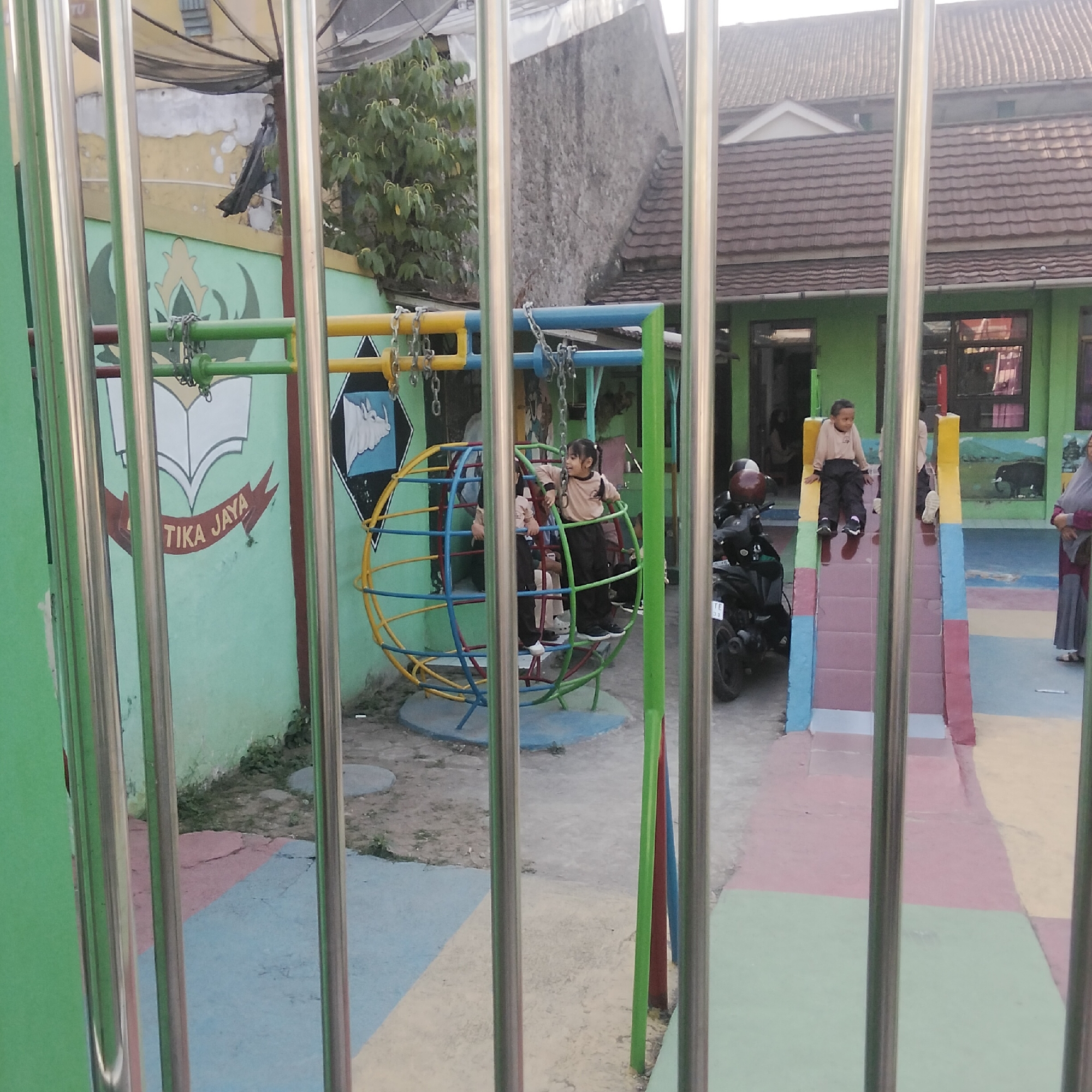 The image shows a playground, likely part of a school, with children playing. In the foreground, a metal fence obscures the view. Through the fence, a colorful play structure resembling a globe is visible, with two children inside it. To the right of the globe, a slide with colorful steps leads down to a multi-colored ground. Another child is seated at the top of the slide. A motorcycle is parked to the left of the slide. The background features a green building with windows and a tiled roof. A mural depicting a rhino is visible on the wall of the building. The ground is paved with colorful squares of red, blue, and yellow. The location is identified as Cisaat, Indonesia, suggesting a school or community center in that area.