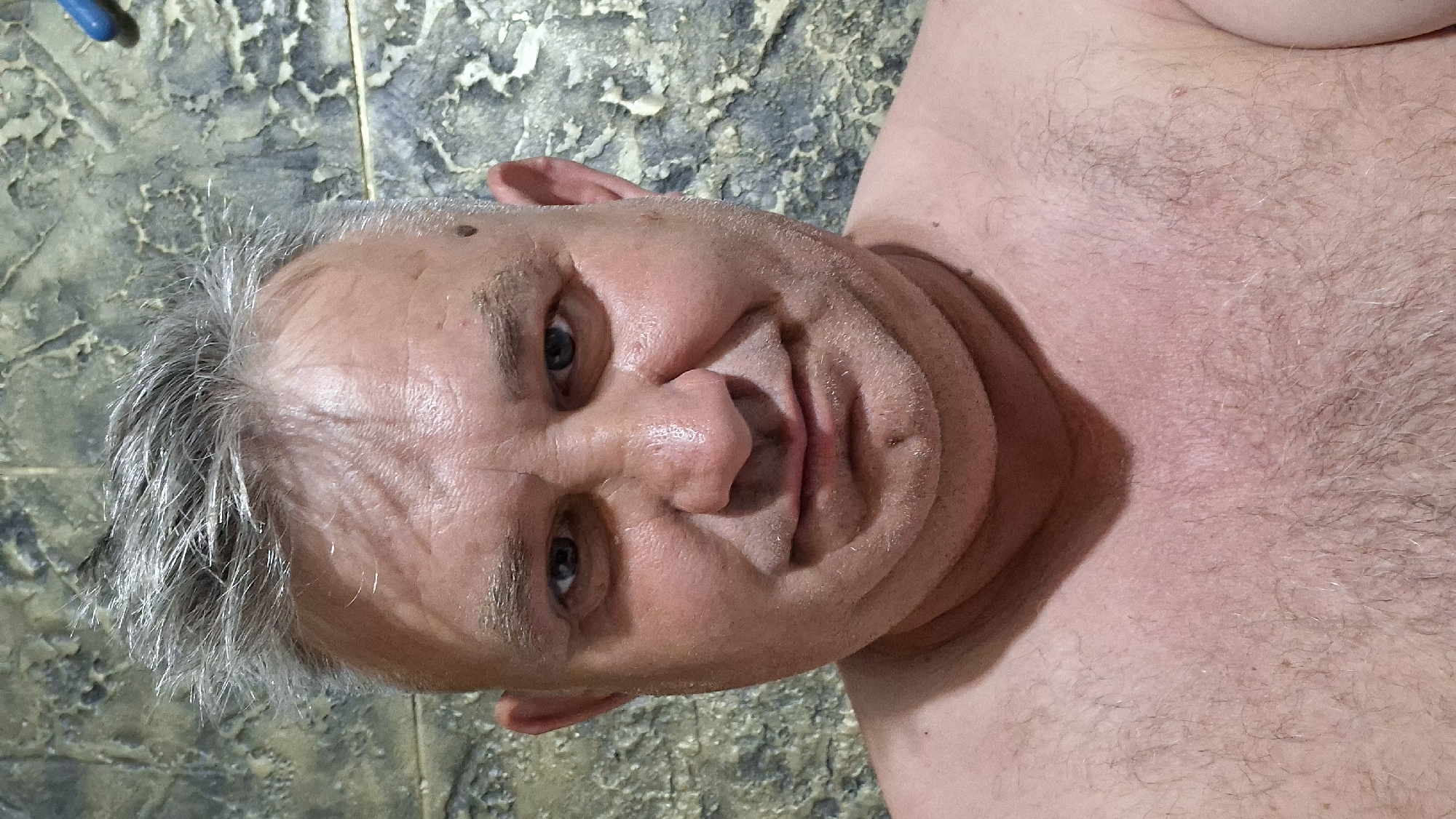 The image is a close-up, head-and-shoulders selfie of a middle-aged man with graying hair. He is looking directly at the camera with a neutral expression. His skin appears flushed, and he has noticeable wrinkles on his forehead. There is a small mole on his left cheek and another on his chin. The background is a textured wall, possibly in a bathroom, with a bluish-gray and green mottled pattern. A small piece of blue plastic is visible in the upper right corner. The man is shirtless, and his chest hair is visible. The lighting is somewhat harsh, creating shadows and highlighting the texture of his skin. There are no other people or objects of significance in the frame. The image does not provide any information about the location in the city of Voinova, Moldova.
