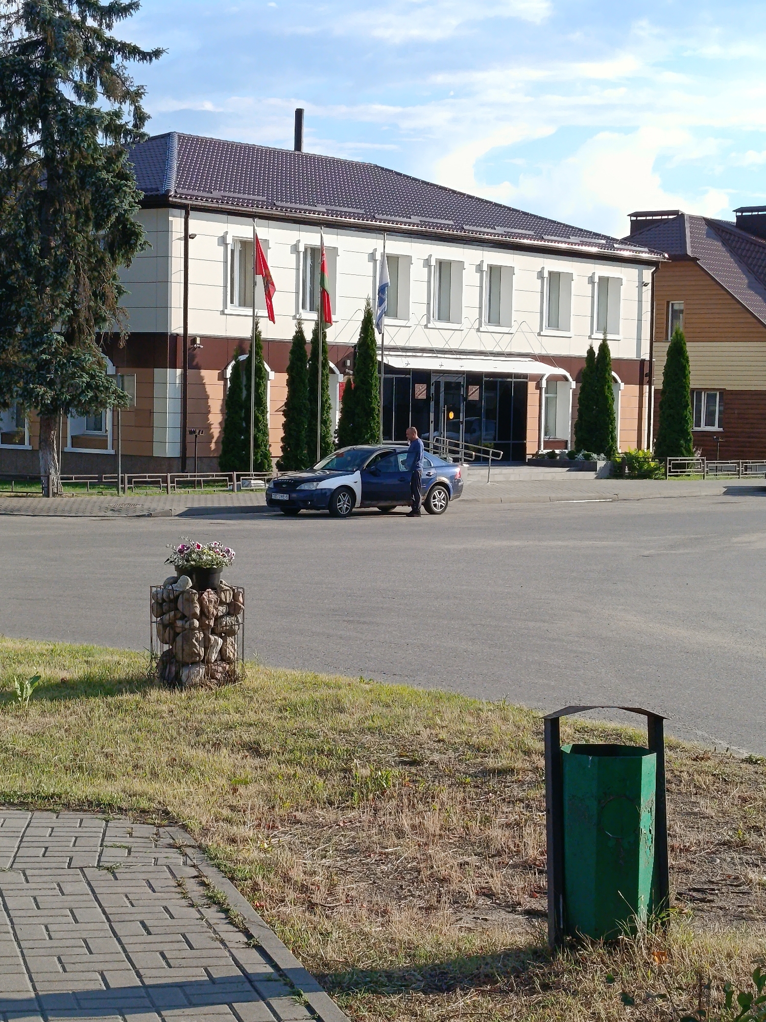 The image depicts an exterior scene in front of a two-story building. The building has a white facade with brown trim and a dark-tiled roof. There are several windows on the second floor and large glass doors on the ground floor. Flags are displayed in front of the building. A dark blue sedan is parked on the asphalt in front of the building, and a person in a blue shirt and dark pants is standing next to the open driver's side door. To the left of the building, there is a large evergreen tree. In the foreground, there is a decorative structure made of stones in a wire cage, topped with a planter containing flowers. A paved walkway with a brick pattern is visible in the lower left corner, and a green trash can is situated on a patch of dry grass to the right. The sky is partly cloudy. The scene is likely located in a public area, possibly a municipal building or business in Смілавіцкі сельскі Савет, Belarus.