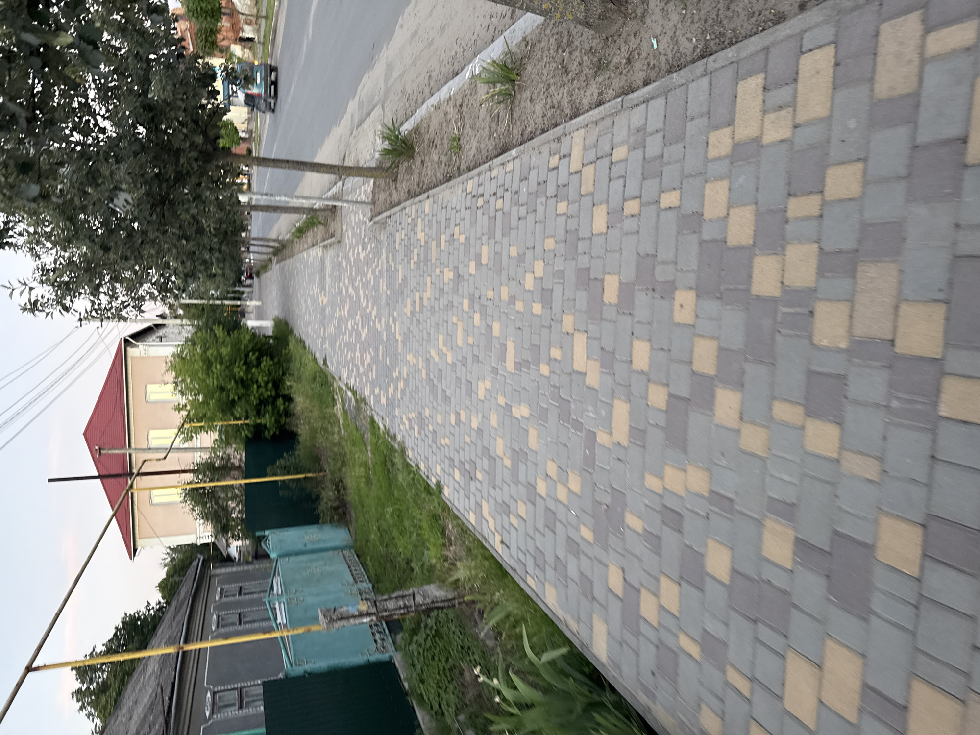 The image depicts a paved sidewalk made of gray and beige interlocking pavers, running alongside a street in Balta, Ukraine. The sidewalk is bordered by a strip of dry soil and sparse vegetation, with several young trees planted along its edge. Beyond the trees, a two-lane asphalt road is visible with a green car driving on it and another car parked further down. On the left side of the sidewalk, a green metal fence encloses a yard with a house visible behind it, characterized by a gray roof and windows. Another house with a red roof and beige facade, also with windows, is situated further down the street. The sky is overcast and appears to be either early morning or late evening. Utility poles and wires are present overhead. The scene is captured from a low-angle perspective, looking down the length of the sidewalk.