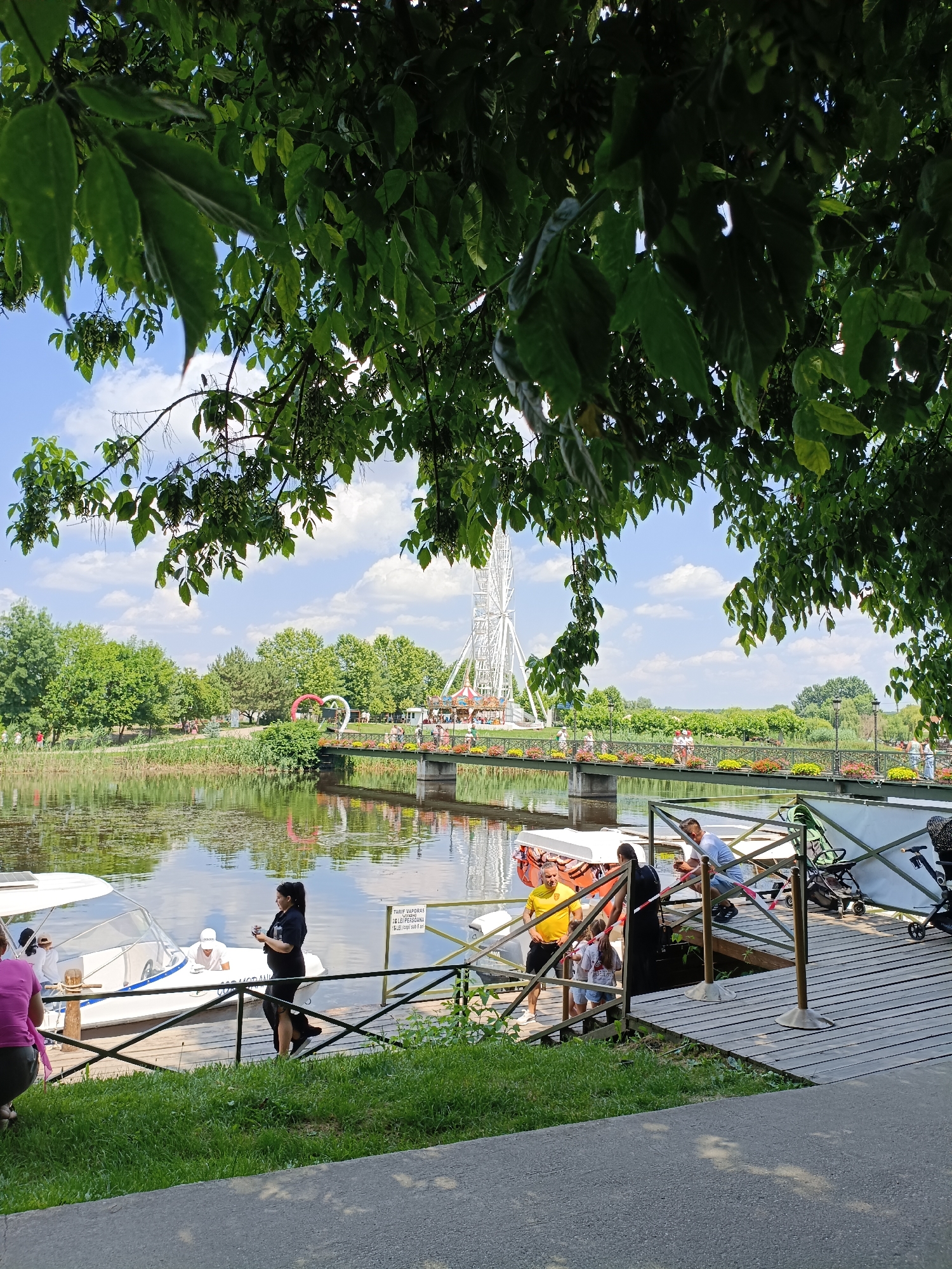 The image shows a bright, sunny day at an amusement park or recreational area next to a lake. In the foreground, a wooden dock juts out into the water, with several small white boats moored. People are standing on the dock, some interacting with the boats, others observing. A woman in a black dress stands on the dock looking at her phone, while a man in a yellow shirt talks to two young girls. Further down the dock, another man in a blue shirt leans against the railing. Green grass lines the edge of the dock, and beyond it, the calm lake reflects the sky and surrounding trees. A bridge spans the lake in the middle ground, adorned with colorful flowers and illuminated heart decorations. Across the bridge, a Ferris wheel and carnival rides are visible, suggesting a lively atmosphere. Lush green trees and a blue sky with scattered white clouds complete the scene. A sign in Romanian, "TARIFE VAPORAS DE LEI PERSOANA 15 LEI (copii sub 6 ani)", is visible on the dock, indicating boat rental prices. The location is the Comana amusement park in Comana, Romania, known for its lake activities and family entertainment.