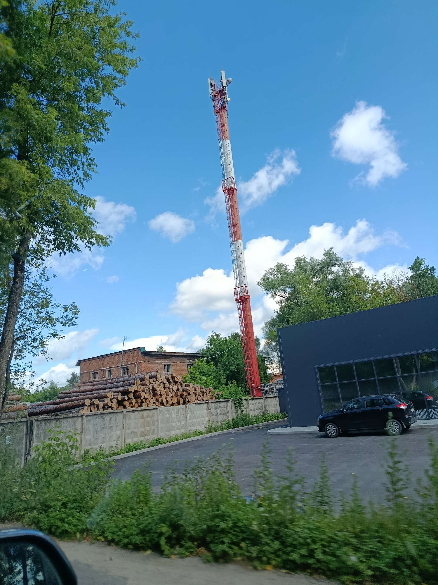A tall red and white cell tower dominates the sky in Orekhovo-Zuyevo, Russia. To the left of the tower, a large tree with green leaves stands tall. In the background, a brick building with several windows is visible, surrounded by a pile of stacked logs. A concrete fence runs along the front of the property. To the right of the tower, a modern dark blue building with large windows is situated. A black car is parked in front of this building, and another car is reflected in the windows. The scene is set against a bright blue sky with scattered white clouds.