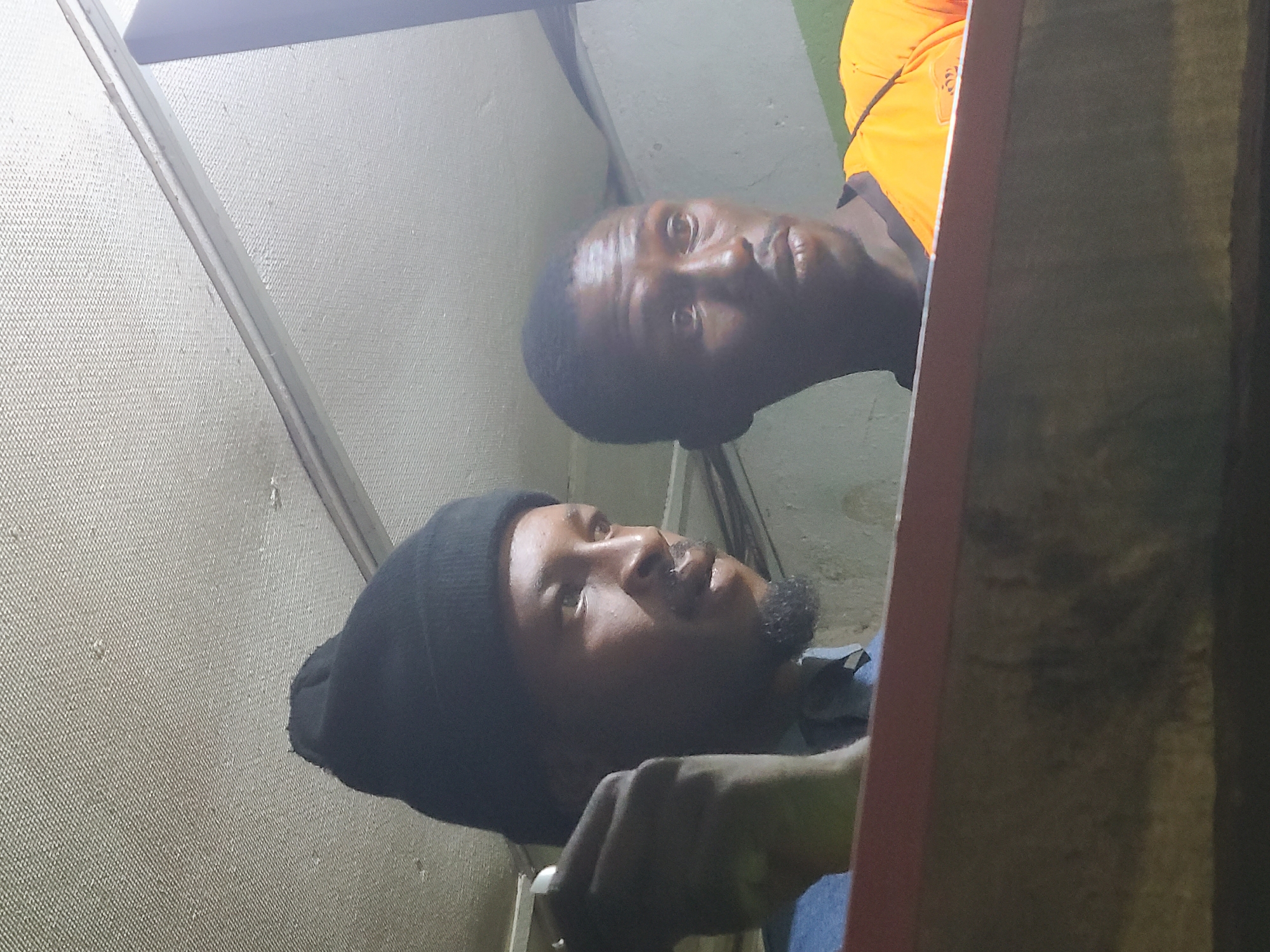 Two African men are indoors. The man in the foreground wears a black beanie and a blue shirt. The man in the background wears an orange shirt. They appear to be looking upwards, perhaps examining the ceiling or upper portion of the wall. The wall surface is a light color with a texture. A red or reddish-brown frame-like object is partially visible on the right side of the image. The location is presumed to be in Jos, Nigeria.