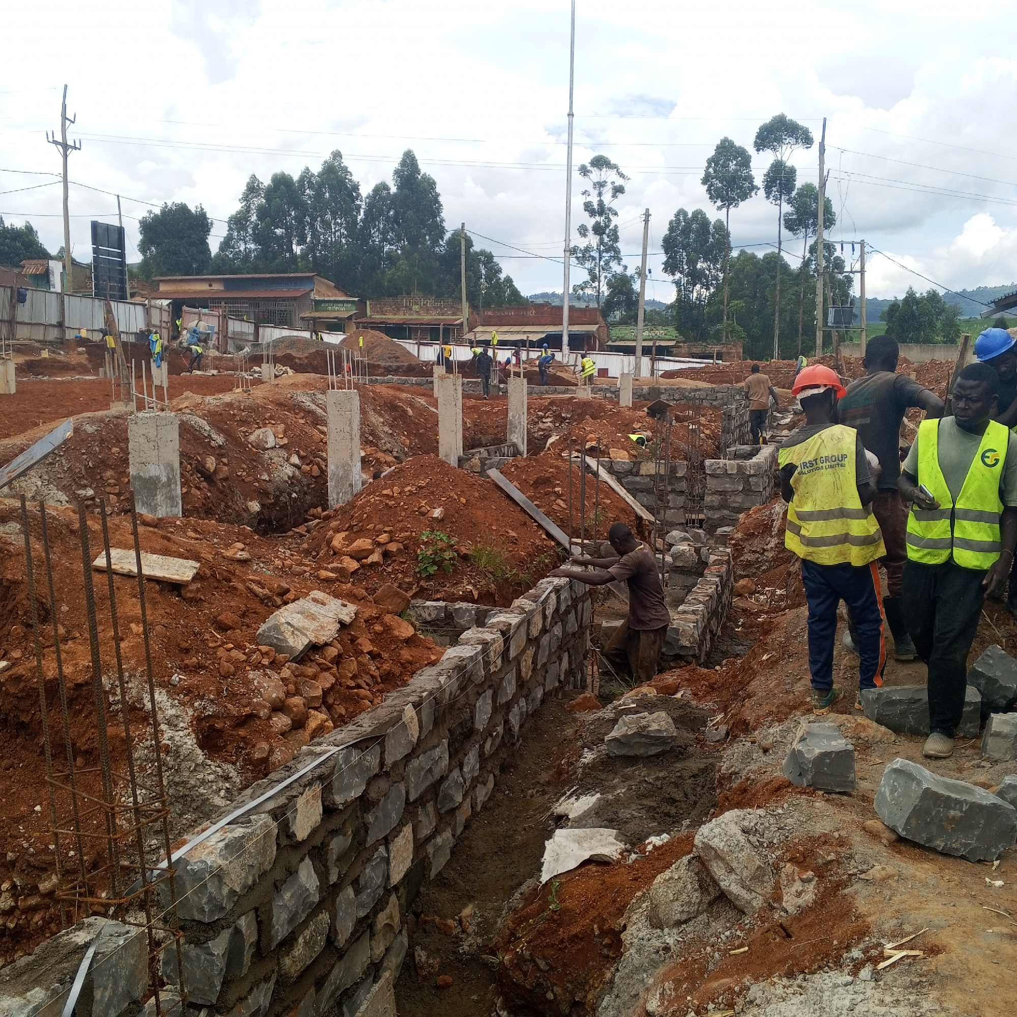 The image depicts a construction site in Borabu Chitago ward, Kenya. Several construction workers are present, some wearing high-visibility vests and hard hats. One worker is laying stones to build a wall, while others appear to be moving materials or supervising the work. The construction site has exposed red soil, partially constructed stone walls, concrete pillars with exposed rebar, and piles of building materials. The surrounding environment includes trees, power lines, and buildings, suggesting the site is located within a populated area.