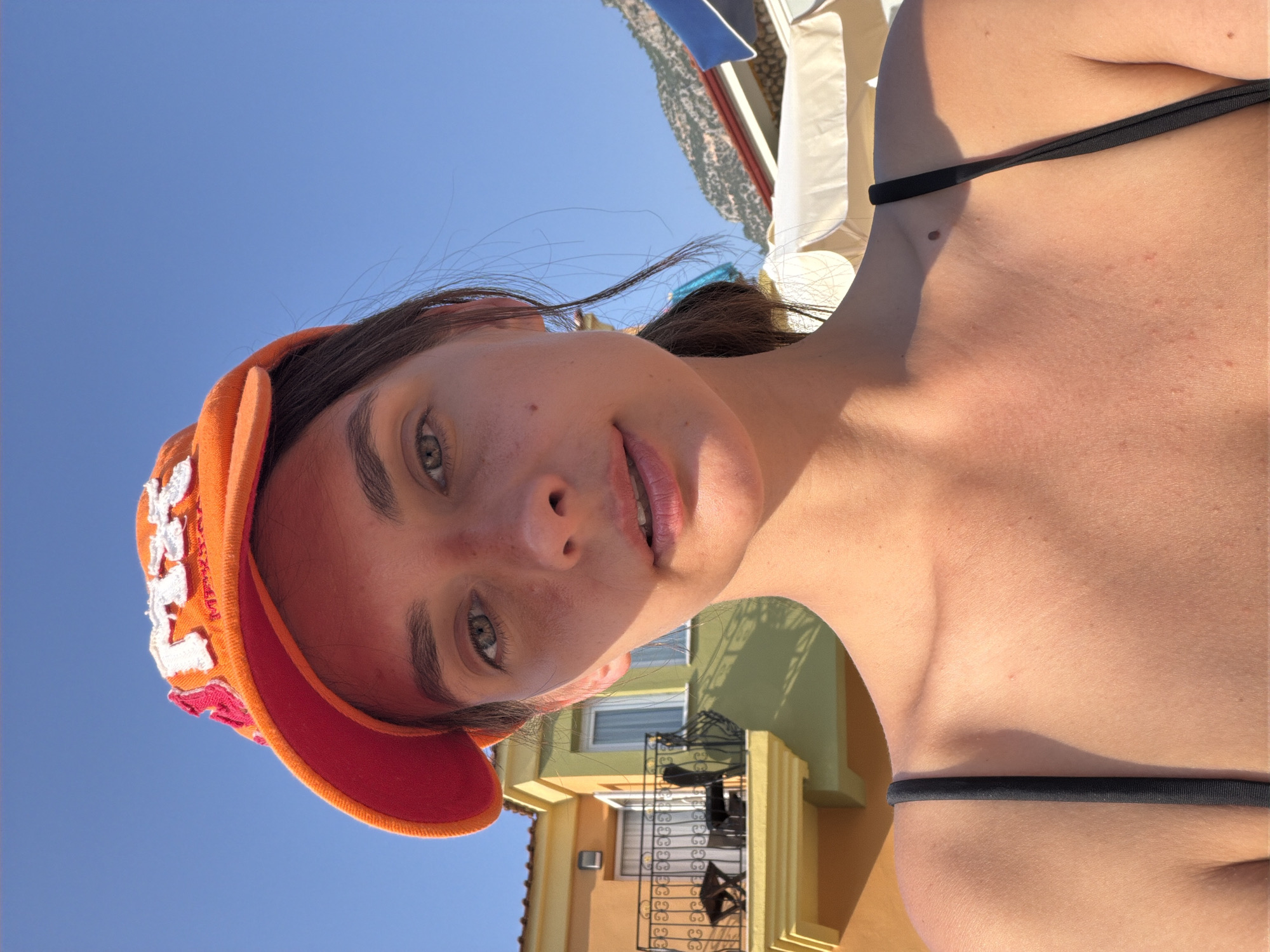 A young woman with brown hair pulled back into a ponytail is taking a selfie outdoors. She is wearing an orange baseball cap with white lettering and a black tank top. Her face is turned slightly towards the camera, and she has a relaxed expression. Behind her, a building with orange and green walls and a balcony with metal railings is visible. The sky is clear and blue, and mountains and beach umbrellas can be seen in the background. The scene appears to be a sunny day in a coastal area, possibly a resort town. The image might have been taken in Fethiye, Turkey, a popular tourist destination known for its beaches and scenery.