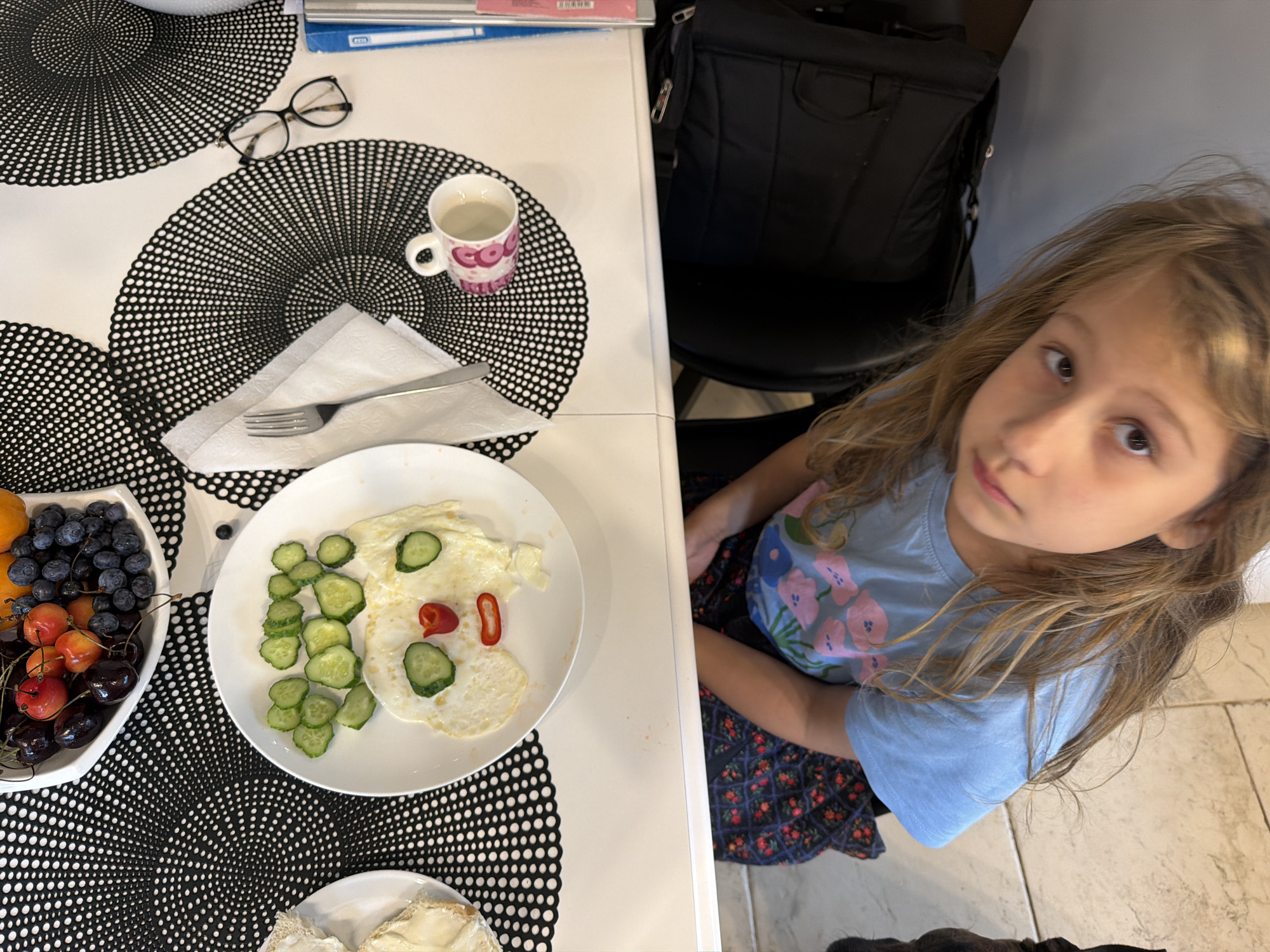 An overhead shot shows a young girl sitting at a white table with black circular placemats. In the foreground, a white plate holds two fried eggs, sliced cucumbers, and two pieces of red pepper, arranged to resemble a face. To the left, a white bowl contains a selection of blueberries, cherries, and apricots. A white mug with a pink and white pattern sits on another placemat, next to a fork and a folded white napkin. A pair of eyeglasses rests on a placemat in the upper left corner. A black backpack is visible behind the girl. The background features a grey wall and tiled flooring. The scene is set in an indoor dining area, possibly a kitchen or dining room.