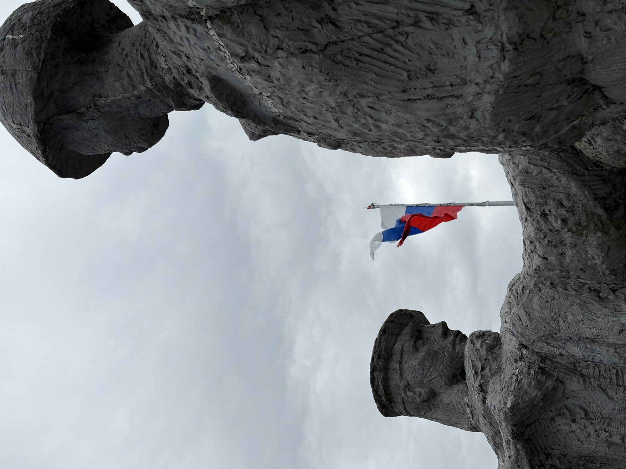 The image shows a low-angle view of a Russian flag waving in the sky, framed by two stone statues of soldiers. The statues are grayscale and have a rough, textured appearance. The sky is overcast and gray. The photo was taken at the Memorial to the Heroic Feat in Wartime in the city of Sochi, Russia.