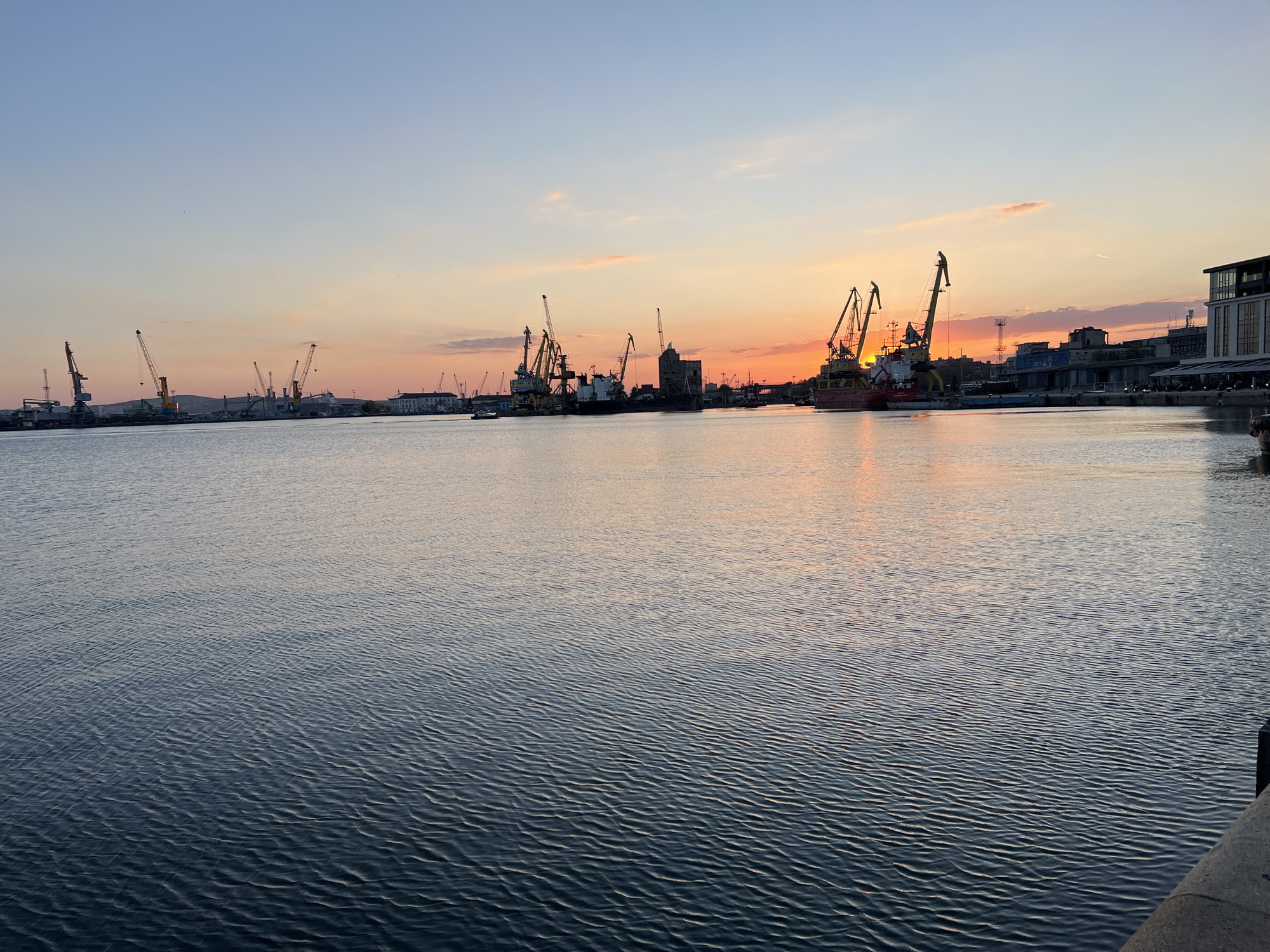 The image depicts the port of Burgas, Bulgaria, during a sunset. The foreground is dominated by the water of the bay, which reflects the colors of the sky. In the background, there are several cranes and industrial structures related to the port operations. A few buildings are visible near the water. The sky is a mix of blue, orange, and pink hues, typical of a sunset.