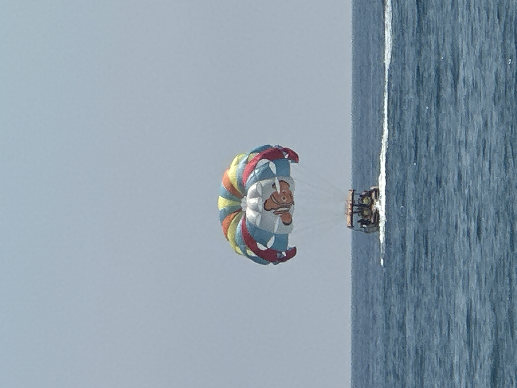 An image shows a boat on the water with a parasail in the sky. The parasail is colorful and has a cartoon fish character on it. The boat is small and has several people on it. The water is blue and there is a clear sky. The scene is set in the city of Boreti, Montenegro.