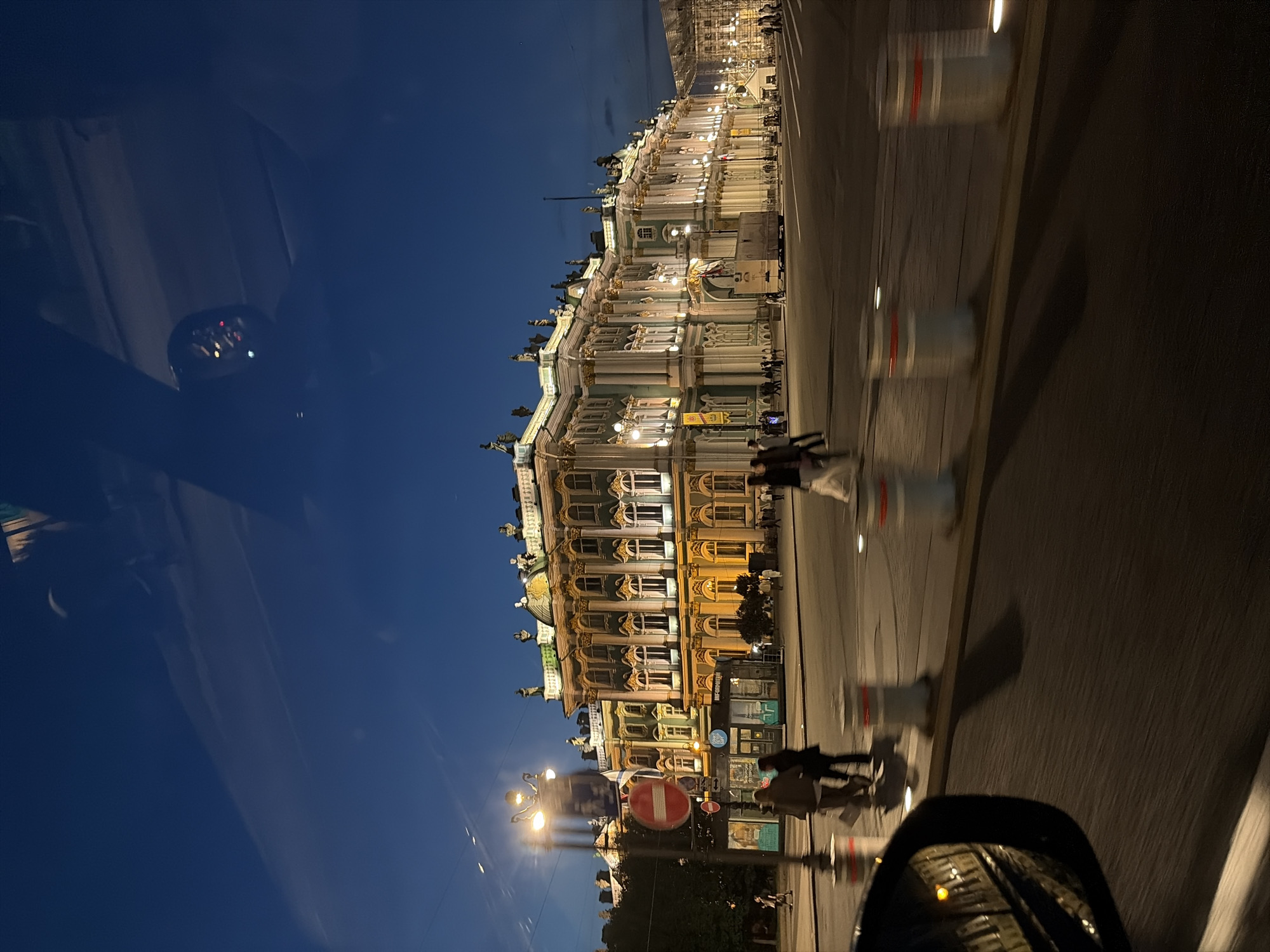 The image depicts the Winter Palace in Saint Petersburg, Russia, at dusk or night. The grand building, illuminated from within, showcases its elaborate architecture with a greenish facade and ornate golden details. Statues adorn the roofline, and numerous windows are lit, casting a warm glow onto the street.
In the foreground, the scene is viewed from inside a vehicle, as evidenced by the car's side mirror reflecting streetlights and the interior of the car. The road in front of the palace is wide, with several pedestrian bollards lining the pavement. Silhouetted figures of people are visible walking on the sidewalk and crossing the street, some appearing to be tourists. The sky is a deep twilight blue. A street lamp with multiple lights stands to the left of the palace, featuring a circular "no entry" sign. A small kiosk with signage in Russian is also present on the sidewalk. The overall impression is of a bustling, historic city square at the end of the day.