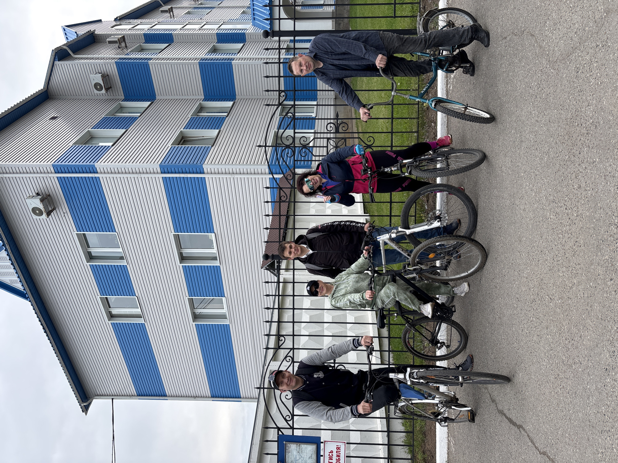 This image, taken in Chaikovsky, Russia, captures five individuals posing with their bicycles in front of a distinct blue and white striped building on an overcast day. The scene unfolds on an asphalt surface, with a black metal fence separating the subjects from a narrow strip of grass and the building beyond.
On the far right, a man in a dark shirt and pants stands beside a light blue bicycle, leaning against the fence. Next to him, a woman sits on a black bicycle, smiling at the camera while holding a Snickers bar. She wears a dark jacket, pink shirt, and black leggings with "PUMA" visible on the leg. Adjacent to her, a man in a dark jacket and jeans stands with his black bicycle. Further left, a young man in a light green tracksuit and a cap sits on a black bicycle, looking towards the viewer. On the far left, another man, wearing a dark jacket with grey sleeves and a baseball cap, is also seated on his bicycle, smiling.
The background is dominated by a multi-story building featuring striking vertical blue and white corrugated metal siding, rectangular windows, and several exterior air conditioning units. Its roof is also blue. The black metal fence, running across the mid-ground, is decorative with ornate scrollwork. The sky is uniformly grey and overcast.
A small sign attached to the fence in the lower-left corner displays Cyrillic text. The words "ОТЧЕТИСЬ" (REPORT) and "С МОБИЛЯ!" (FROM MOBILE!) are clearly visible, suggesting it's an instruction or advertisement related to mobile reporting. The overall mood of the image is casual and friendly, with the individuals appearing relaxed as they pose for the camera.