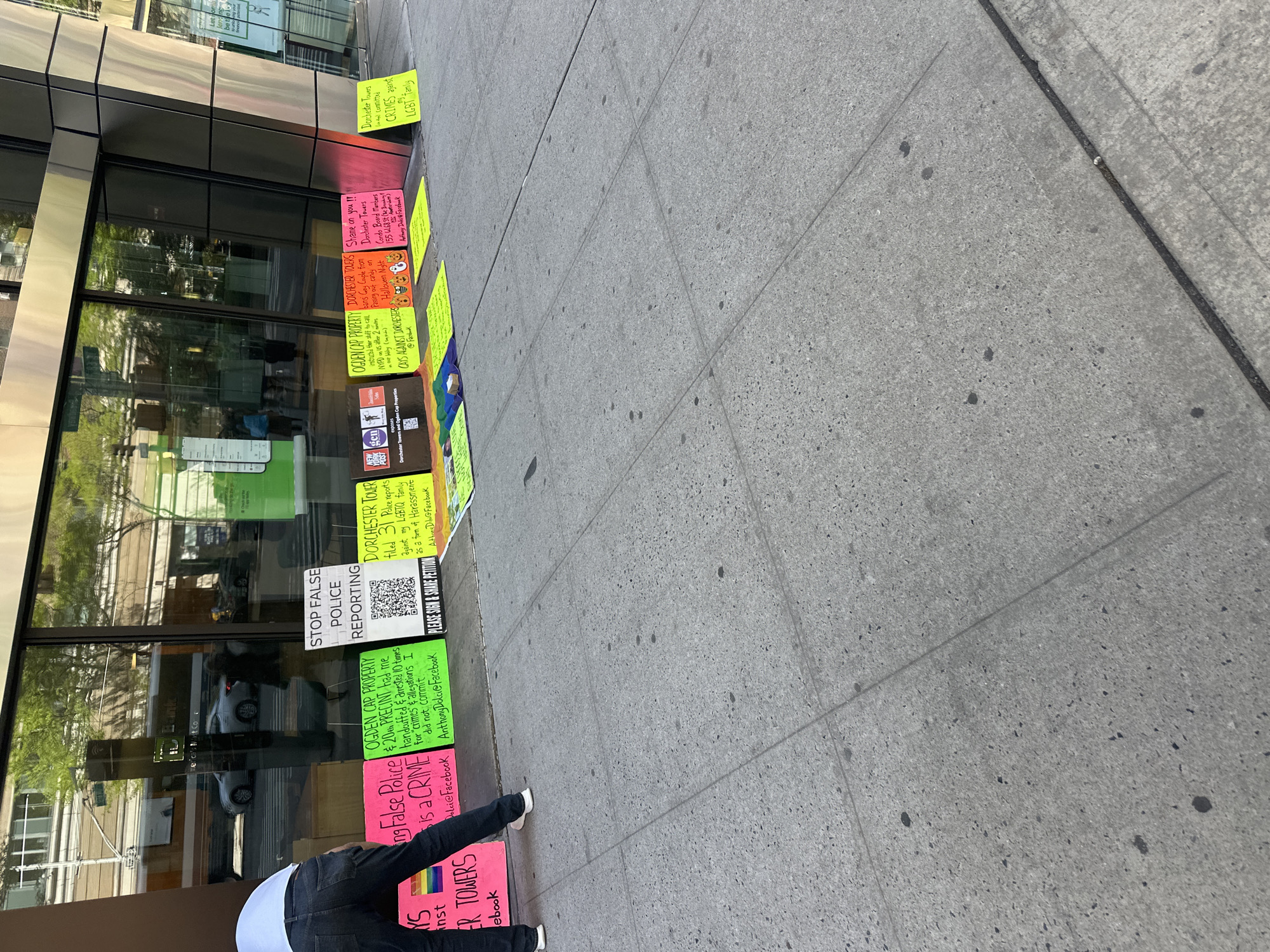 A person is placing or adjusting a series of signs on the sidewalk in front of a building in New York City, United States of America. The signs address concerns related to Dorchester Towers and Ogden Cap Properties, alleging false police reporting, harassment, and crimes against the LGBTQ community. One sign specifically mentions the 20th precinct. Some signs include Facebook handles and QR codes, urging viewers to sign a petition. The building has reflective windows.