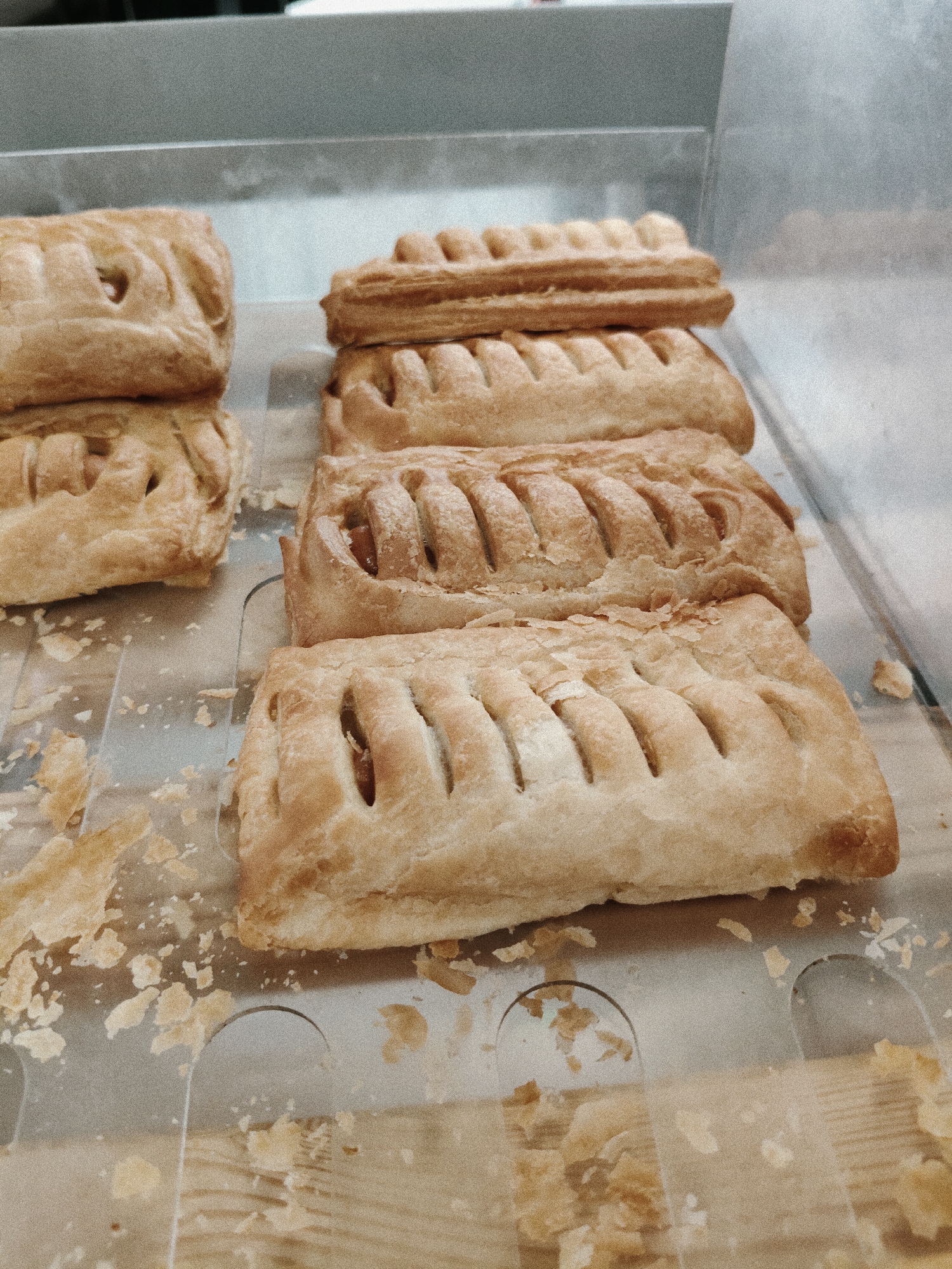 This media file features a close-up shot of several golden-brown, rectangular pastries arranged on a tray. The pastries appear to be filled, with visible layers of flaky dough and some filling peeking through slitted openings on top. Scattered around the pastries are small flakes of pastry, suggesting they are fresh or have been handled. The tray is placed on a light-colored surface, possibly a counter or a display case, with some wooden elements visible beneath a translucent protective layer. The background is blurred, showing a metallic or gray surface, indicating an indoor setting like a bakery or a food service establishment. The lighting is soft, casting a warm glow on the pastries. There are no people or text visible in the image. The scene evokes a sense of deliciousness and freshness, typical of baked goods ready for sale or consumption.